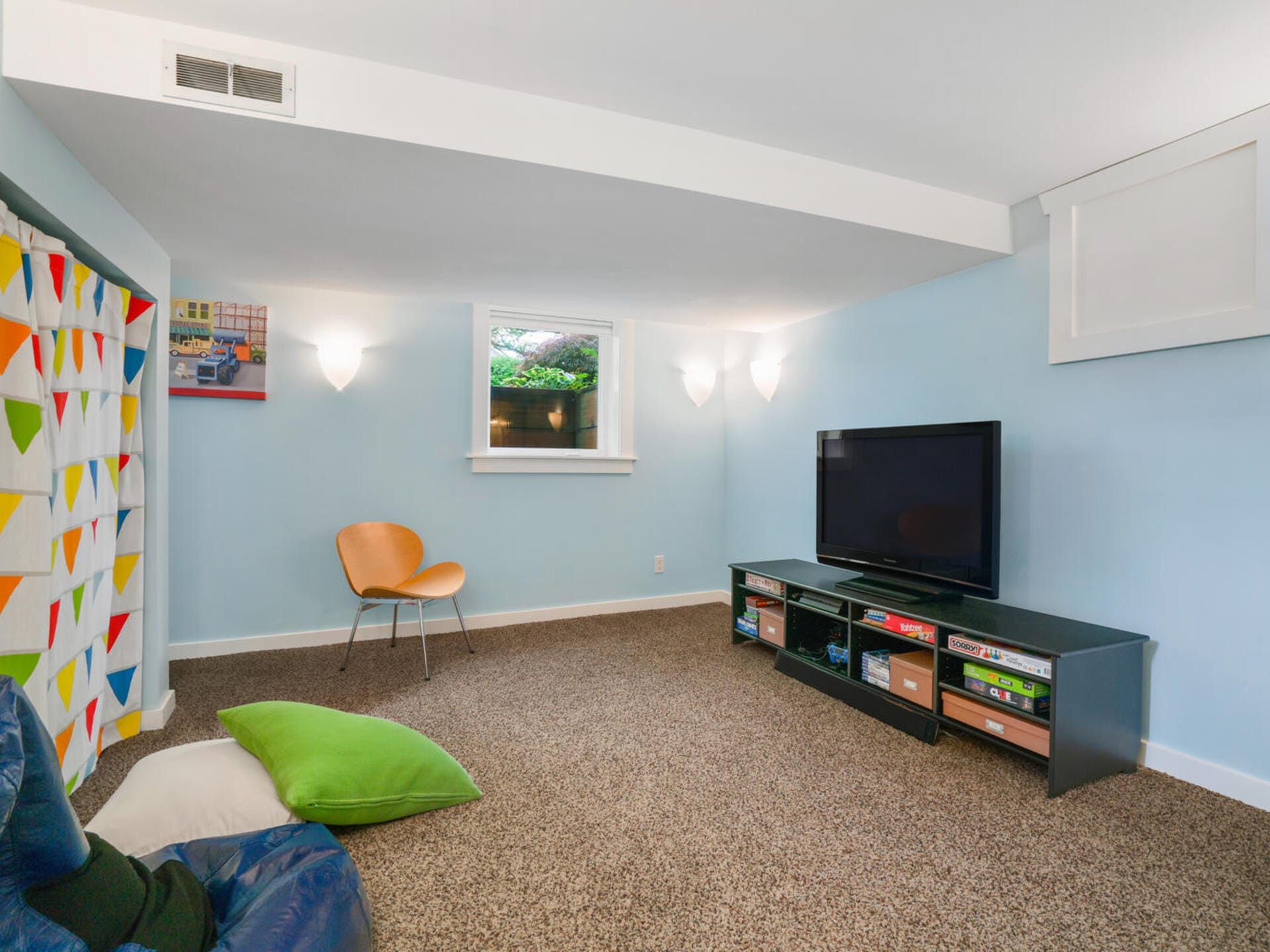 A cozy basement room with light blue walls and carpeted floor. It features a large TV on a stand with shelves, a wooden chair, and a colorful curtain. Soft lighting from wall sconces creates a warm atmosphere.