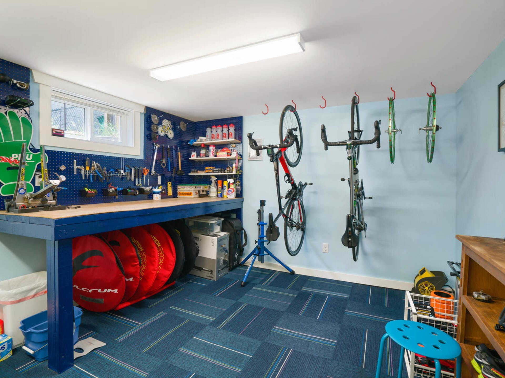 A well-organized bike workshop with blue walls and floor. Bicycles hang on wall hooks, and a workbench holds tools, supplies, and spare parts. A variety of cycling gear, including helmets and wheels, are neatly arranged around the room.