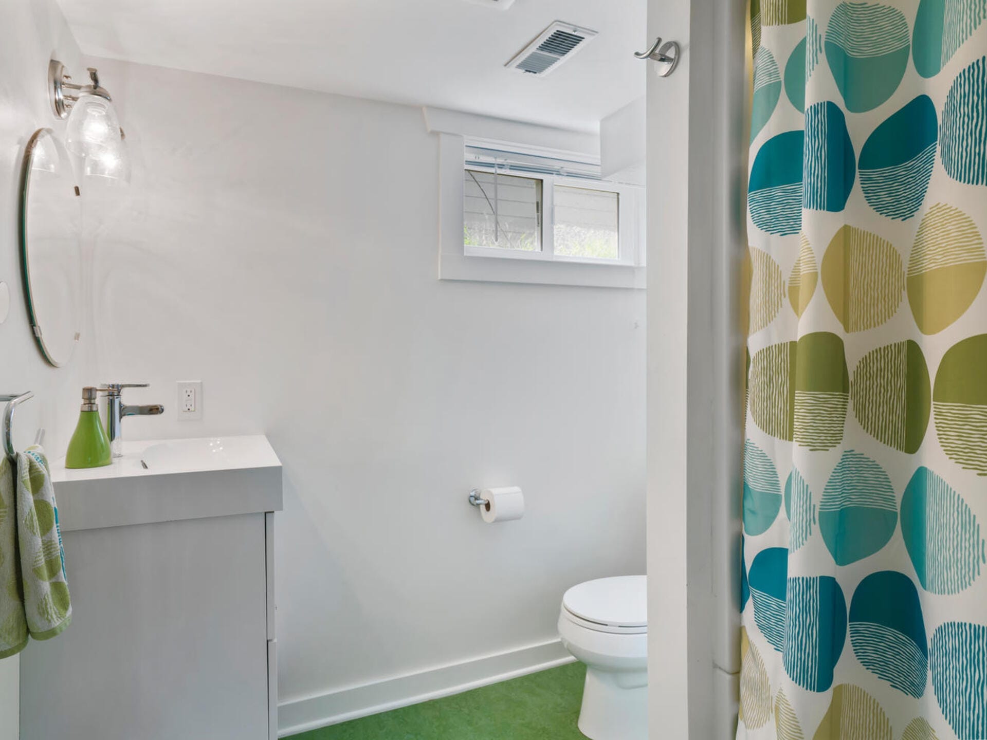 A clean, modern bathroom with green flooring. There is a white sink with a green soap dispenser and hand towel, a round mirror, a toilet, and a shower with a curtain featuring blue and green oval patterns. A small window lets in natural light.