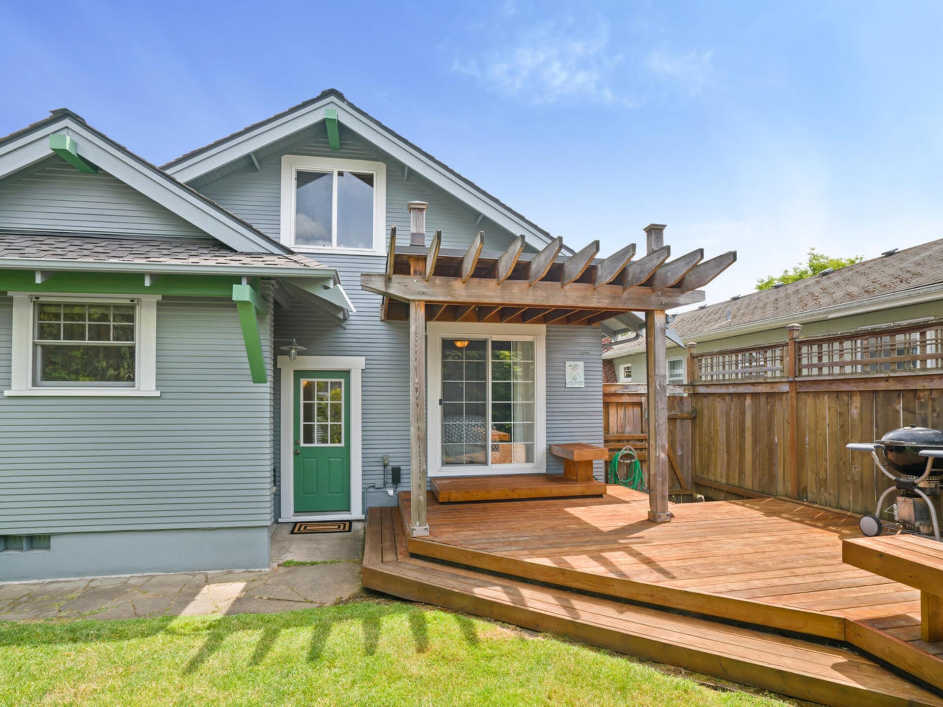 Gray house with a green door, sloping roof, and a wooden pergola on a deck. The yard is grassy, and a barbecue grill sits at the edge of the deck. Theres a wooden fence on the right, and the sky is clear and blue.
