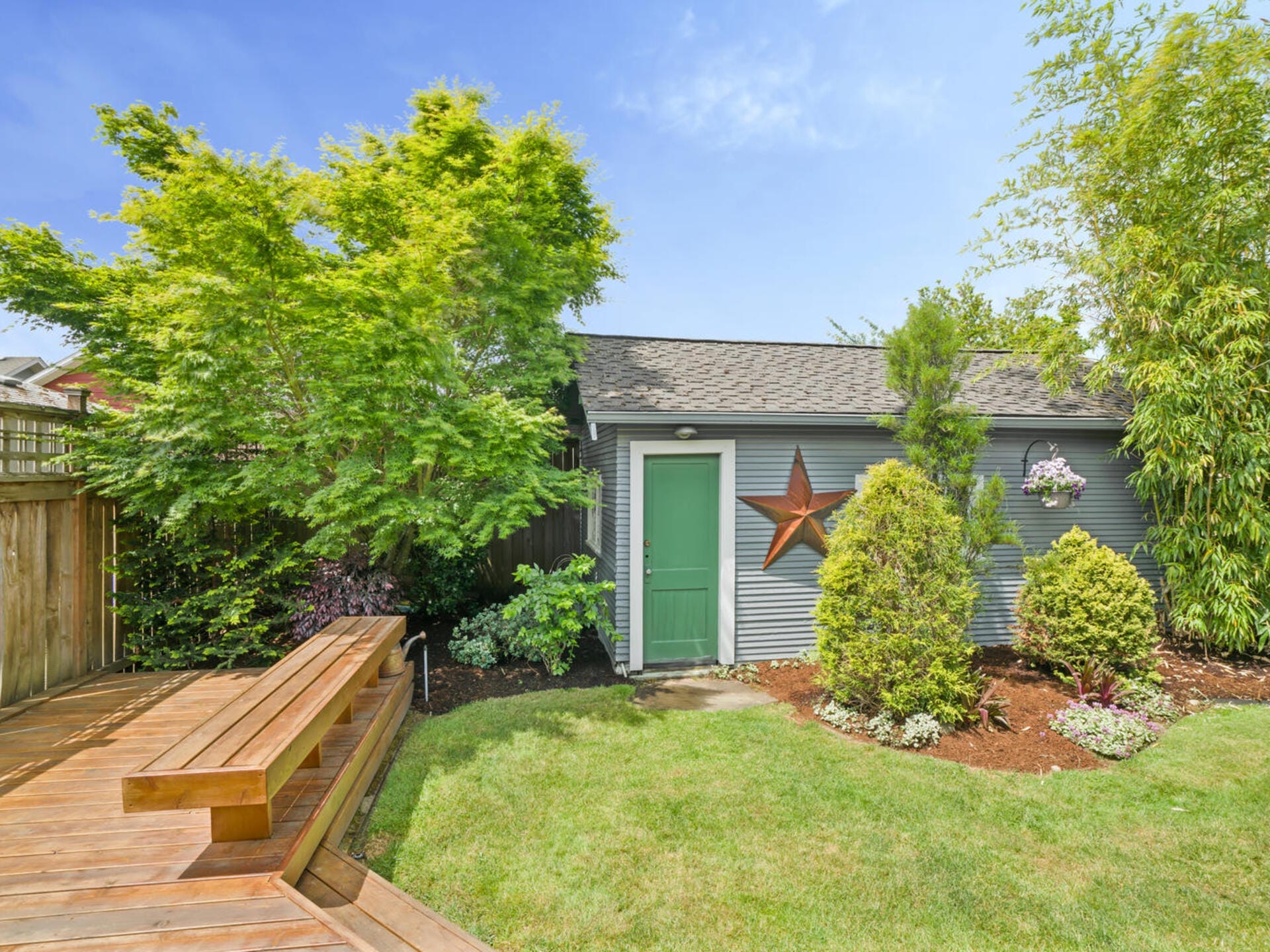 A backyard with green grass and a wooden deck featuring built-in benches. A small gray shed with a green door is adorned with a star decoration and flower baskets, surrounded by trees and shrubs under a clear blue sky.