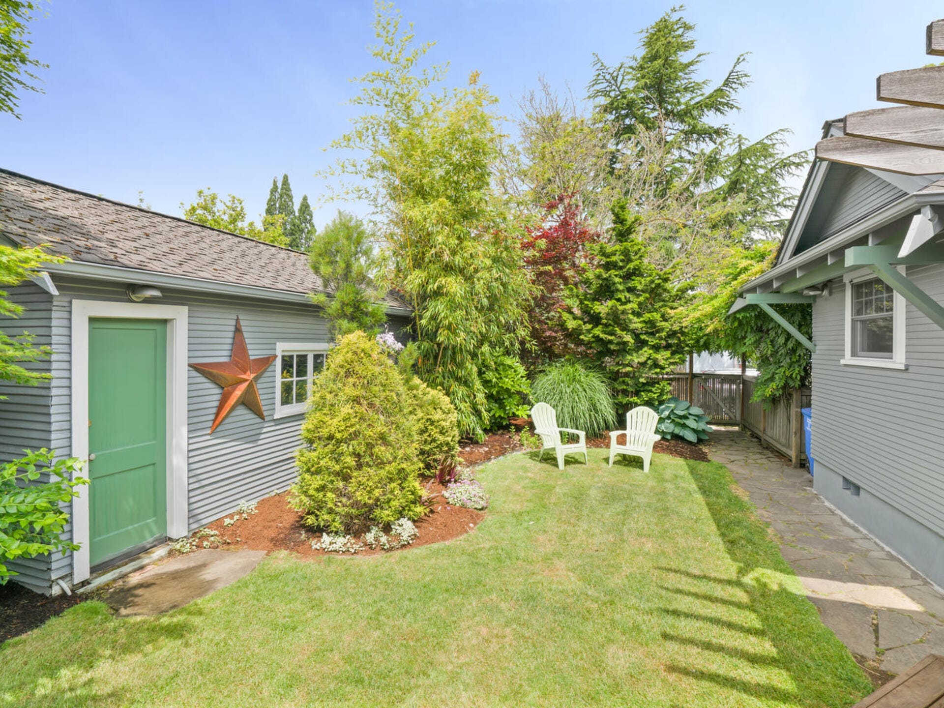 A backyard features a green door with a star decoration on a light blue house. There are two white Adirondack chairs on a small patio surrounded by lush greenery, including trees and shrubs, under a clear blue sky.