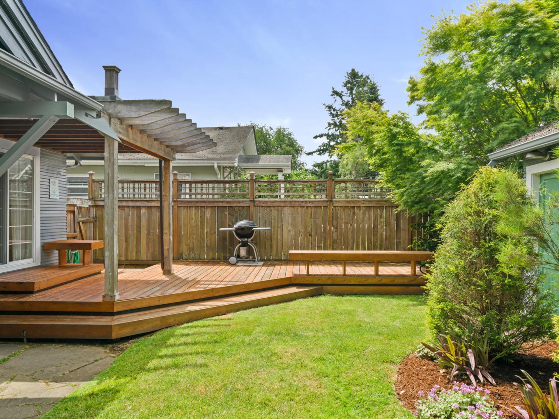 A backyard with a wooden deck and pergola, a grill, neatly trimmed grass, and landscaped plants. A wooden fence encloses the area, and trees provide shade. A clear blue sky is visible above.