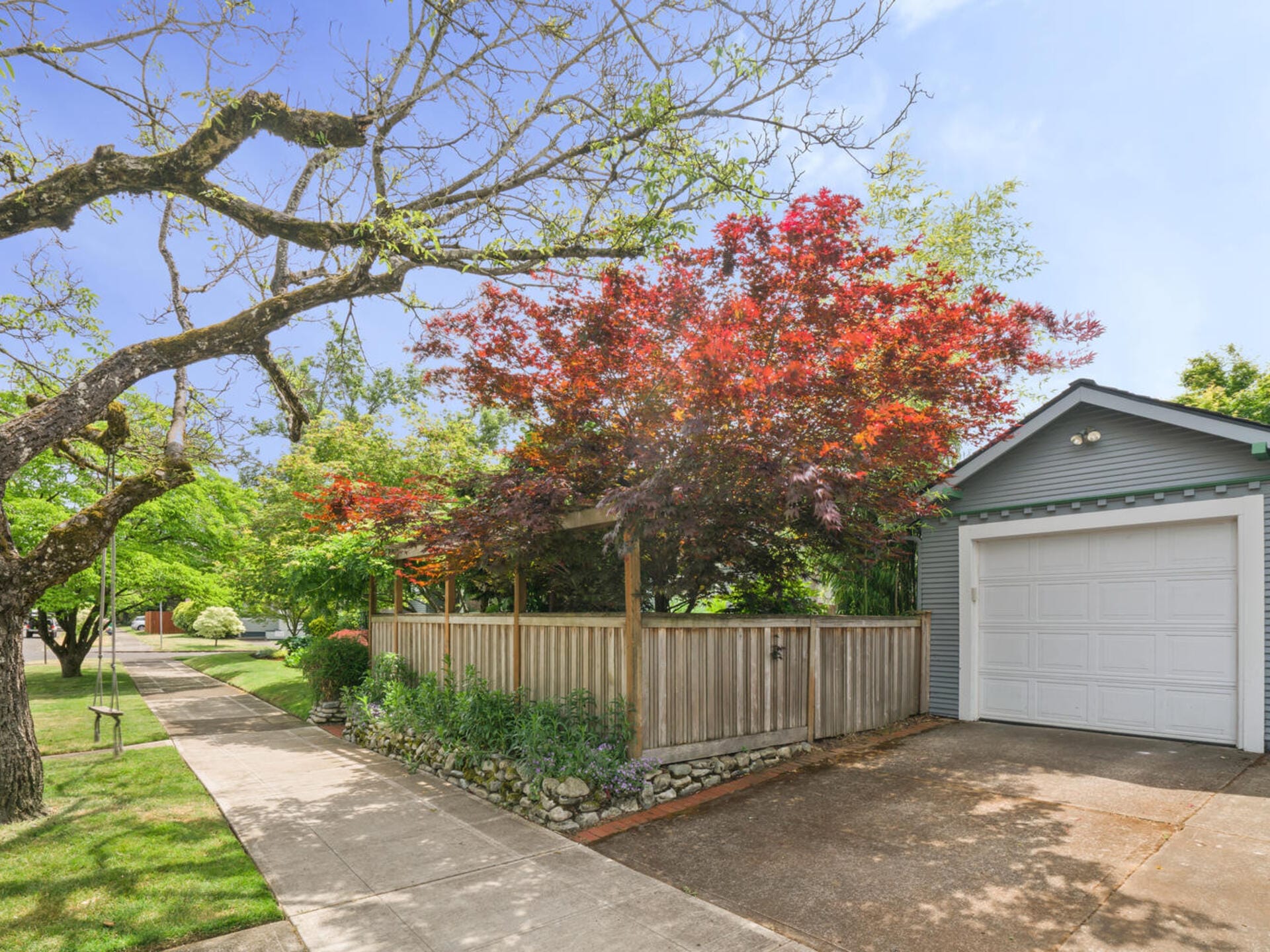 Street view of a quiet neighborhood featuring a house with a garage and a red-leafed tree. A wooden fence borders the property next to a sidewalk, and a tree swing hangs from a branch over the lawn. The sky is clear and blue.