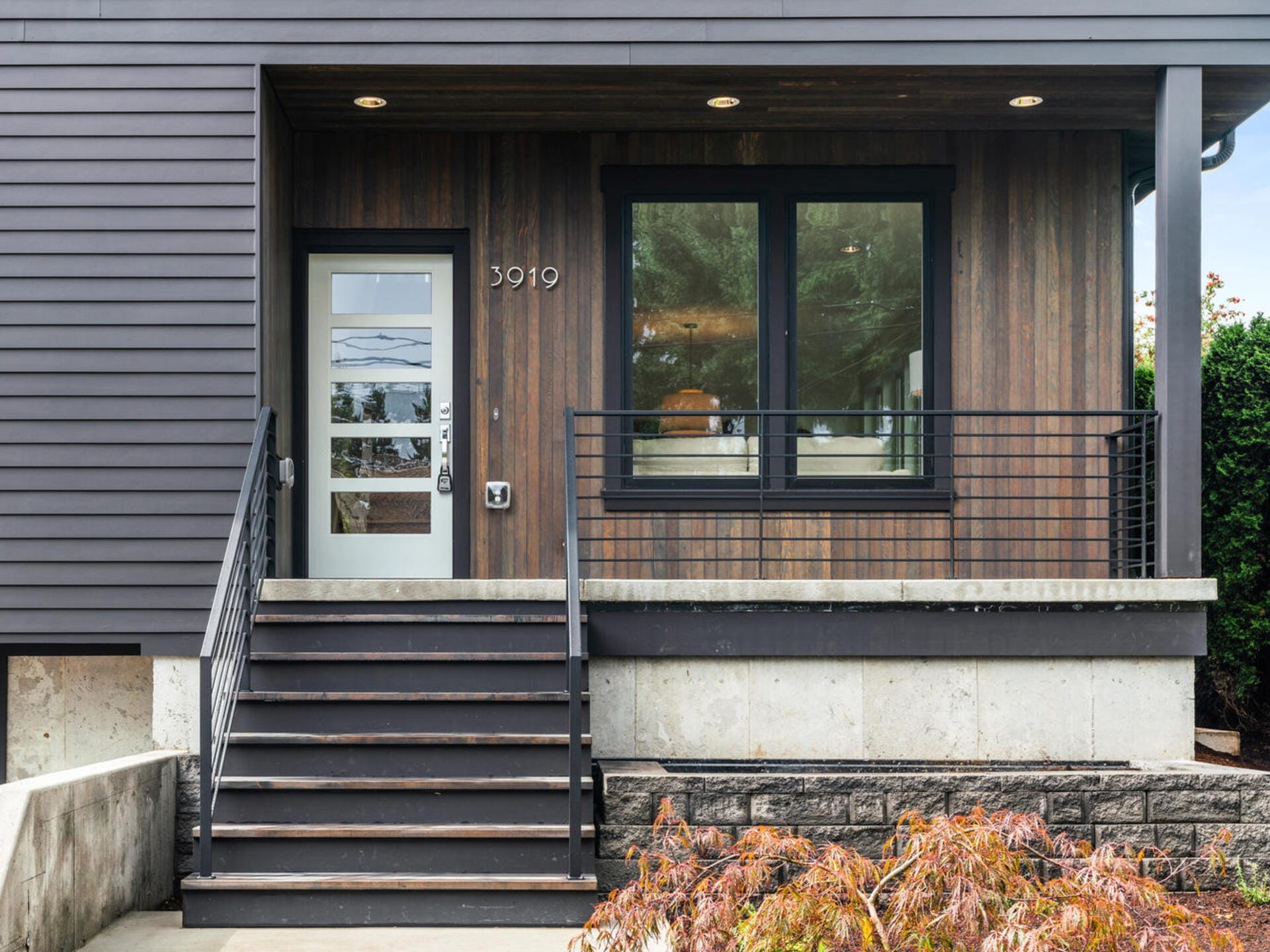 Modern front entrance of a house with horizontal wooden siding and large windows. A set of black metal-railed stairs leads to a door with geometric designs. The house number 3919 is displayed beside the door. Autumn shrubs are in the foreground.