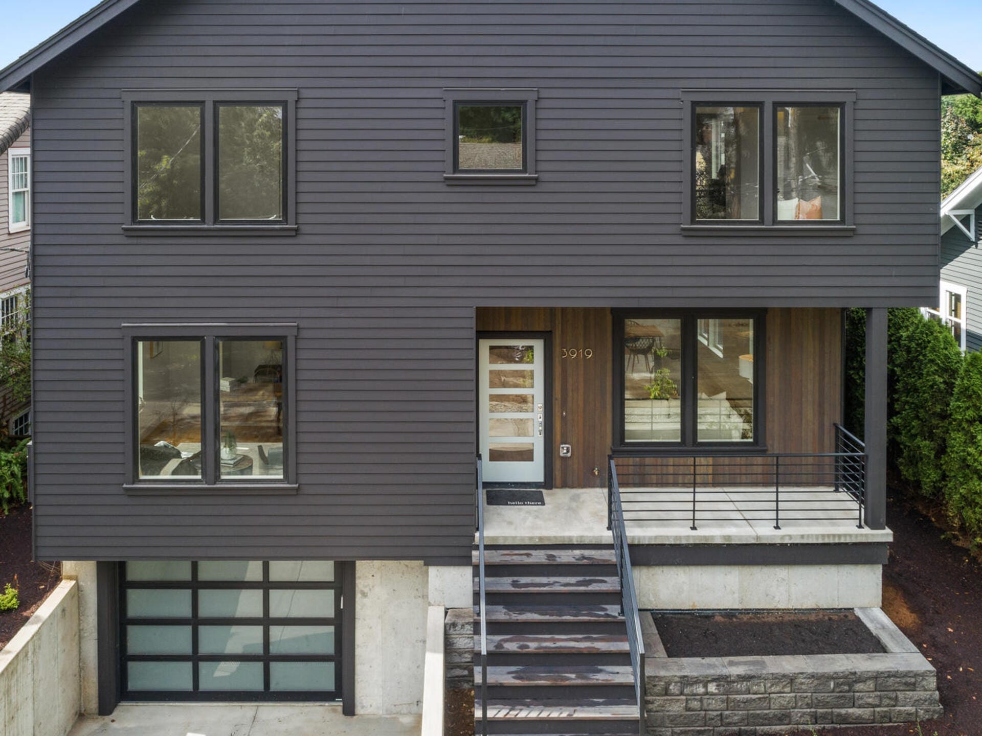 Modern two-story house with dark grey siding, large windows, and a flat front lawn. The entrance features a set of stone steps leading to a porch with a railing. The garage door is glass-paneled.