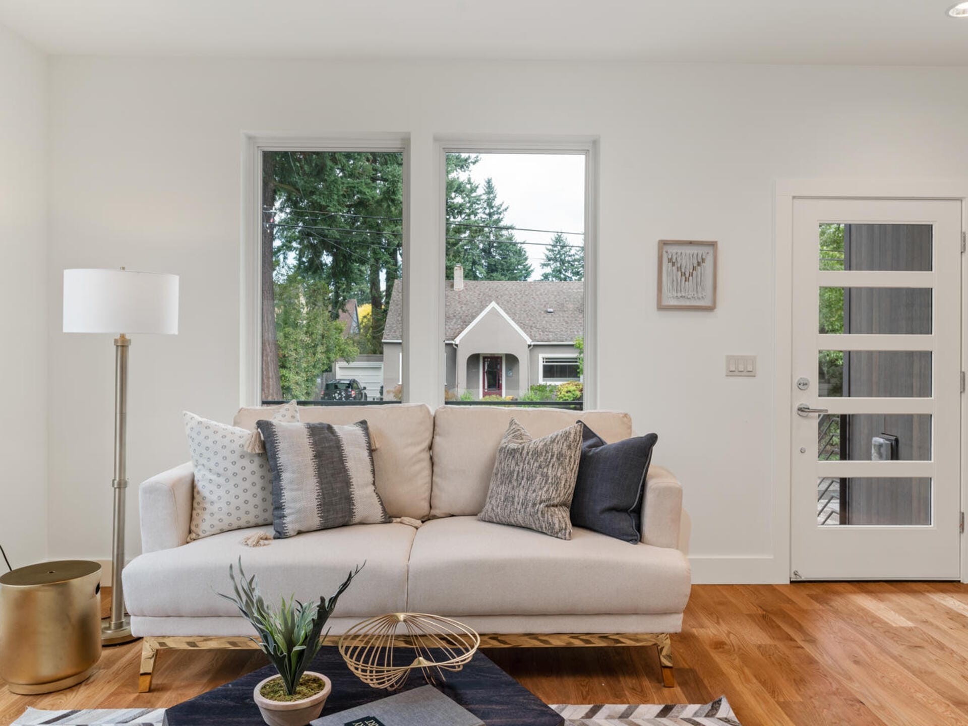 A modern living room features a beige sofa with several pillows, a gold side table with a potted plant, a floor lamp, and a geometric rug. Large windows and a glass-paneled door let in natural light, providing a view of the trees and house outside.