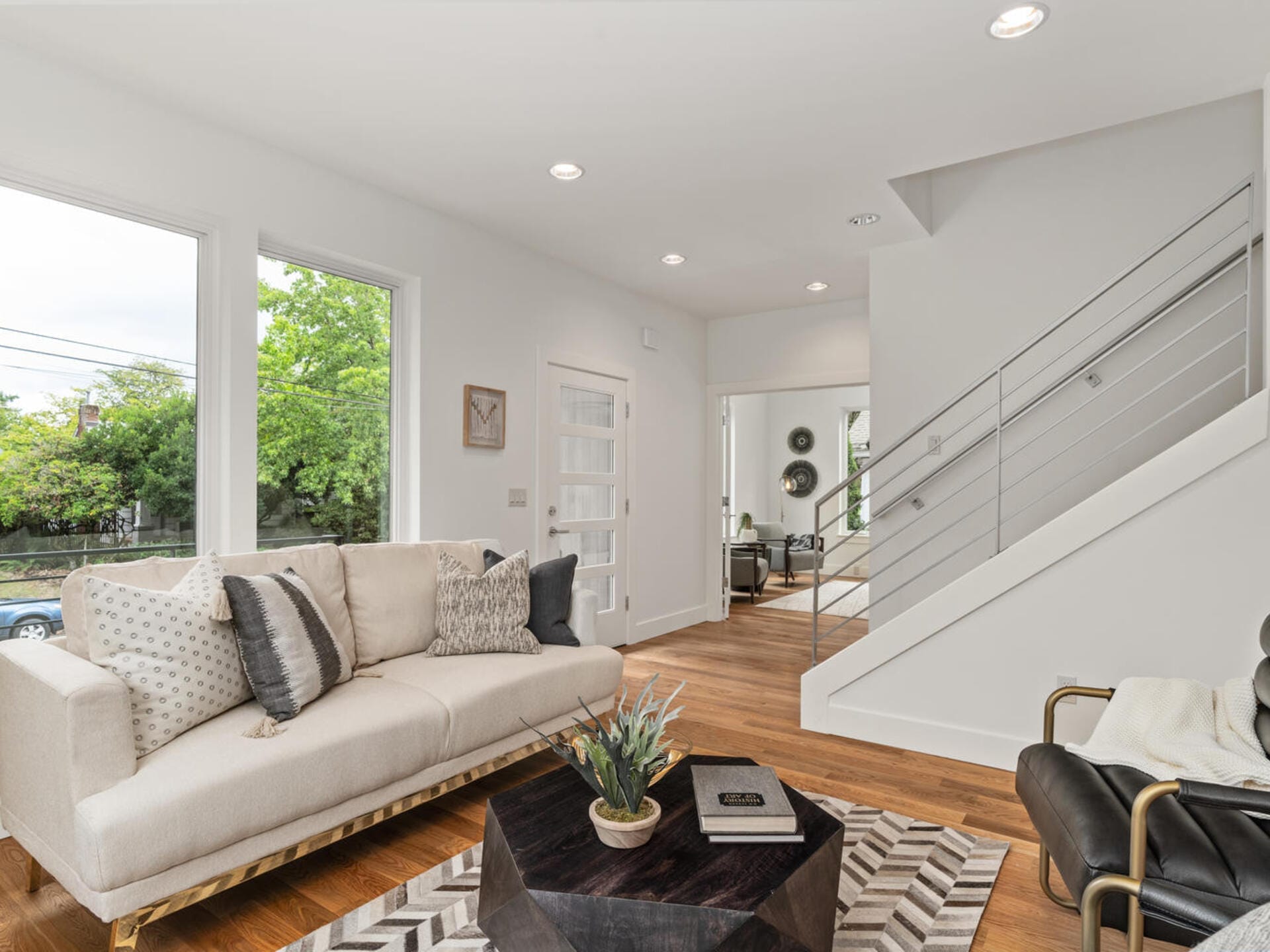 A modern living room with a beige sofa adorned with pillows, a geometric-patterned rug, and a black coffee table with books and a plant. Theres a staircase with a metal railing and large windows that let in natural light.
