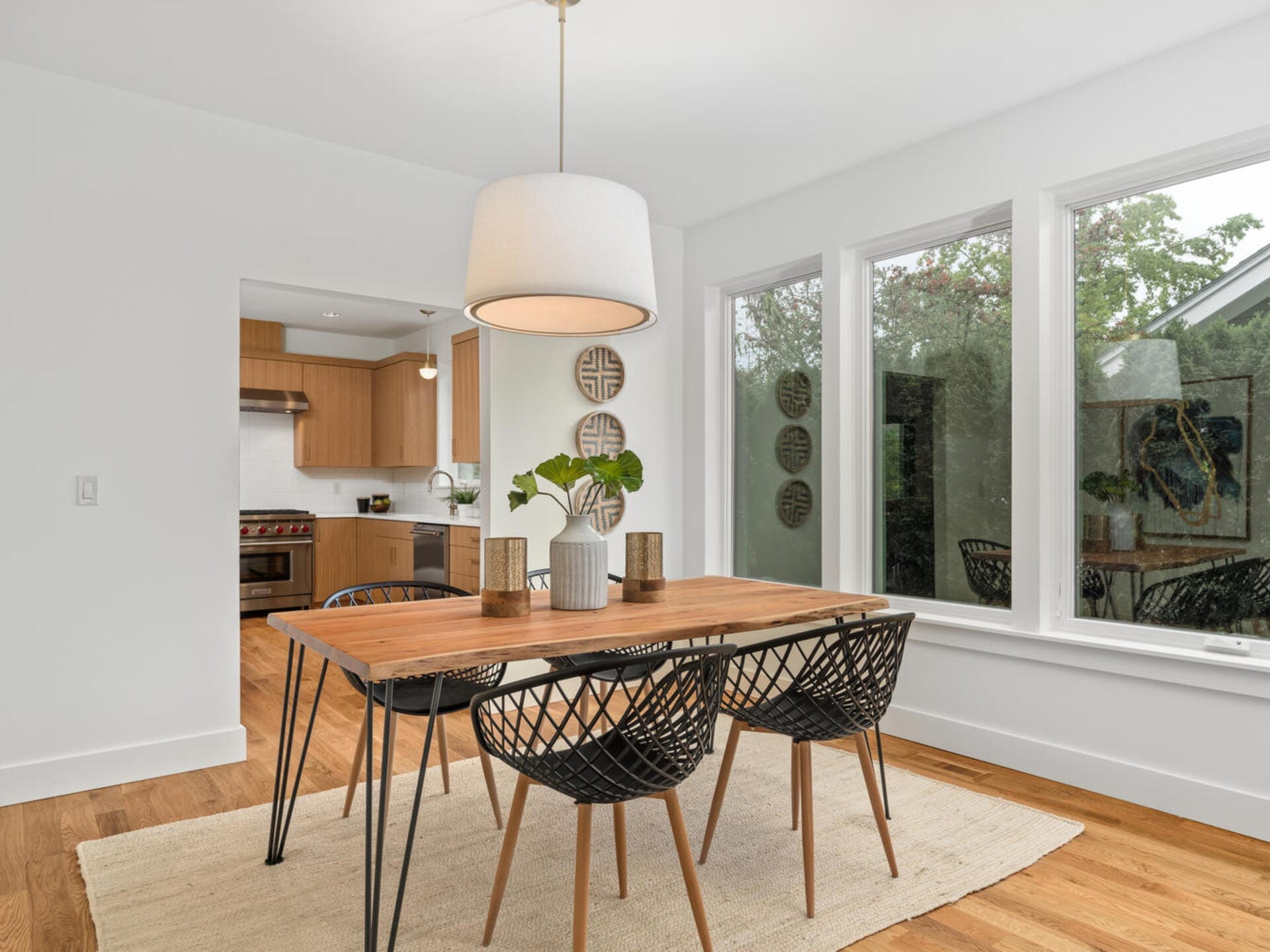 A modern dining room with a wooden table, four black woven chairs, and a large white pendant light. The room features wooden floors, a rug, and large windows providing a view of greenery. The kitchen is visible in the background.