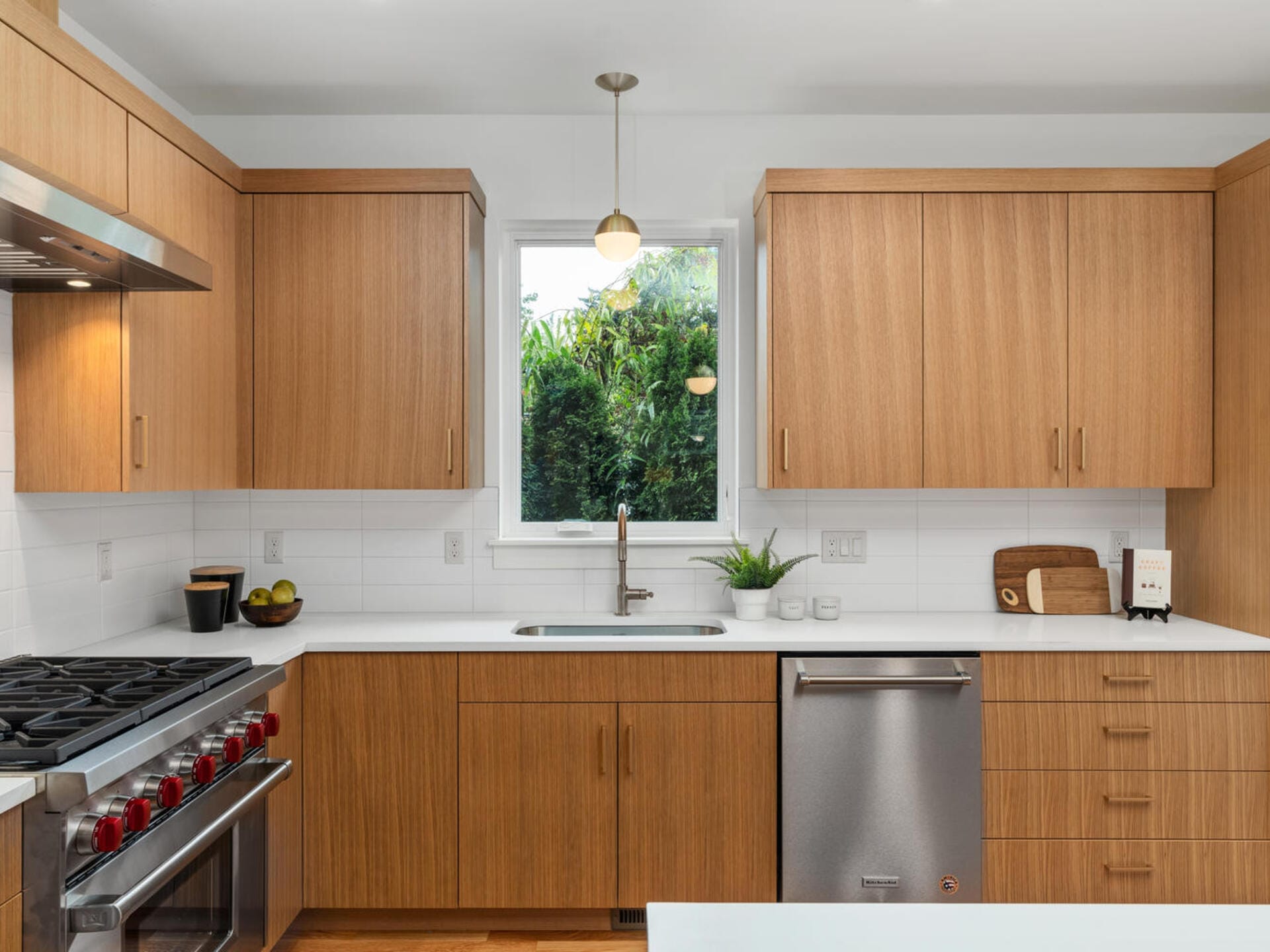 A modern kitchen with wood cabinets, a stainless steel dishwasher, and a gas stove. A window above the sink shows greenery outside. The countertop holds a fruit bowl, potted plant, and cutting board. A pendant light hangs from the ceiling.