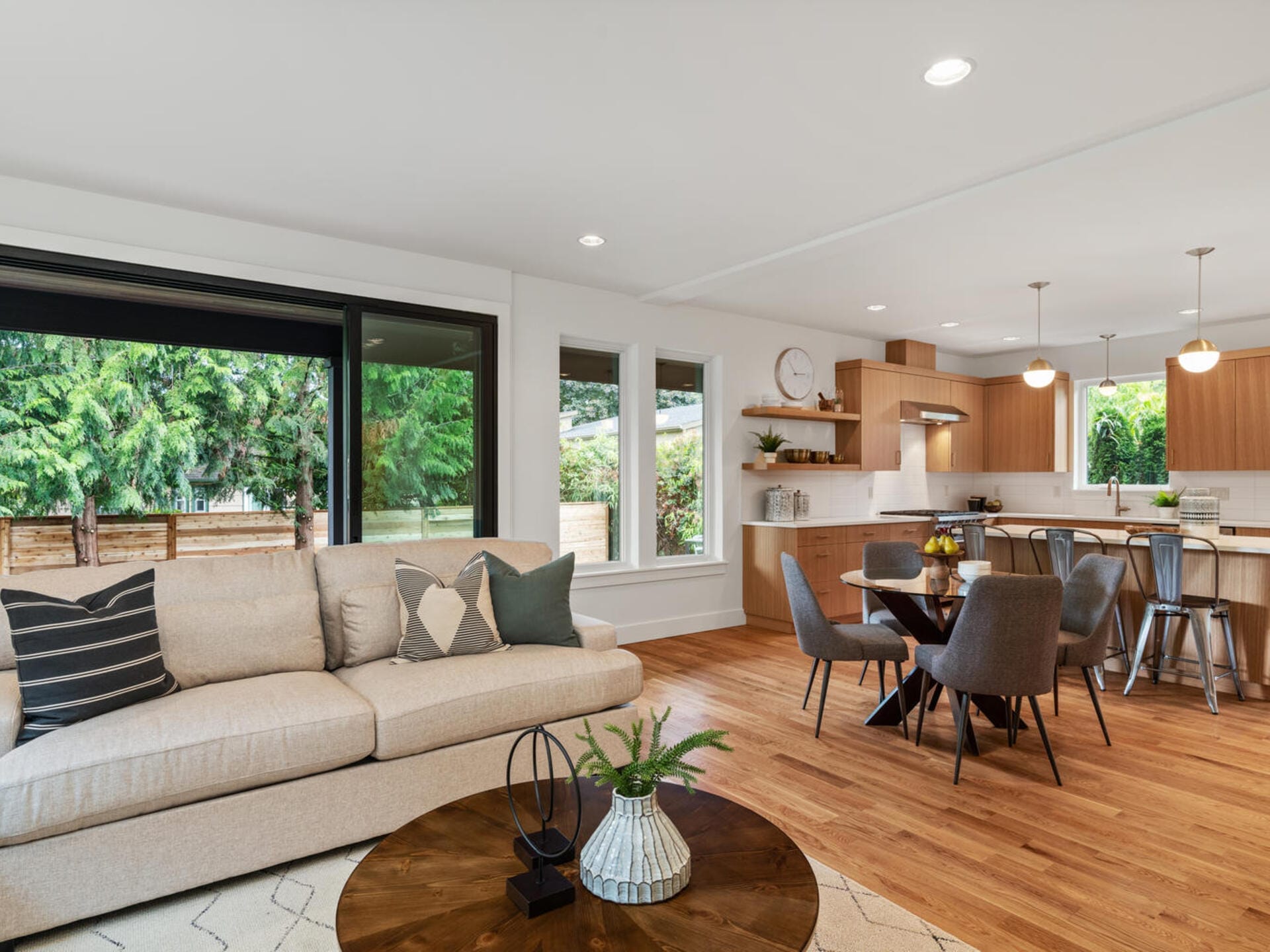 A modern open-plan living and kitchen area with hardwood floors. The space features a beige sofa, round wooden coffee table, and gray dining chairs around a circular table. Large windows showcase a lush green garden outside. Pendant lights hang above the kitchen island.