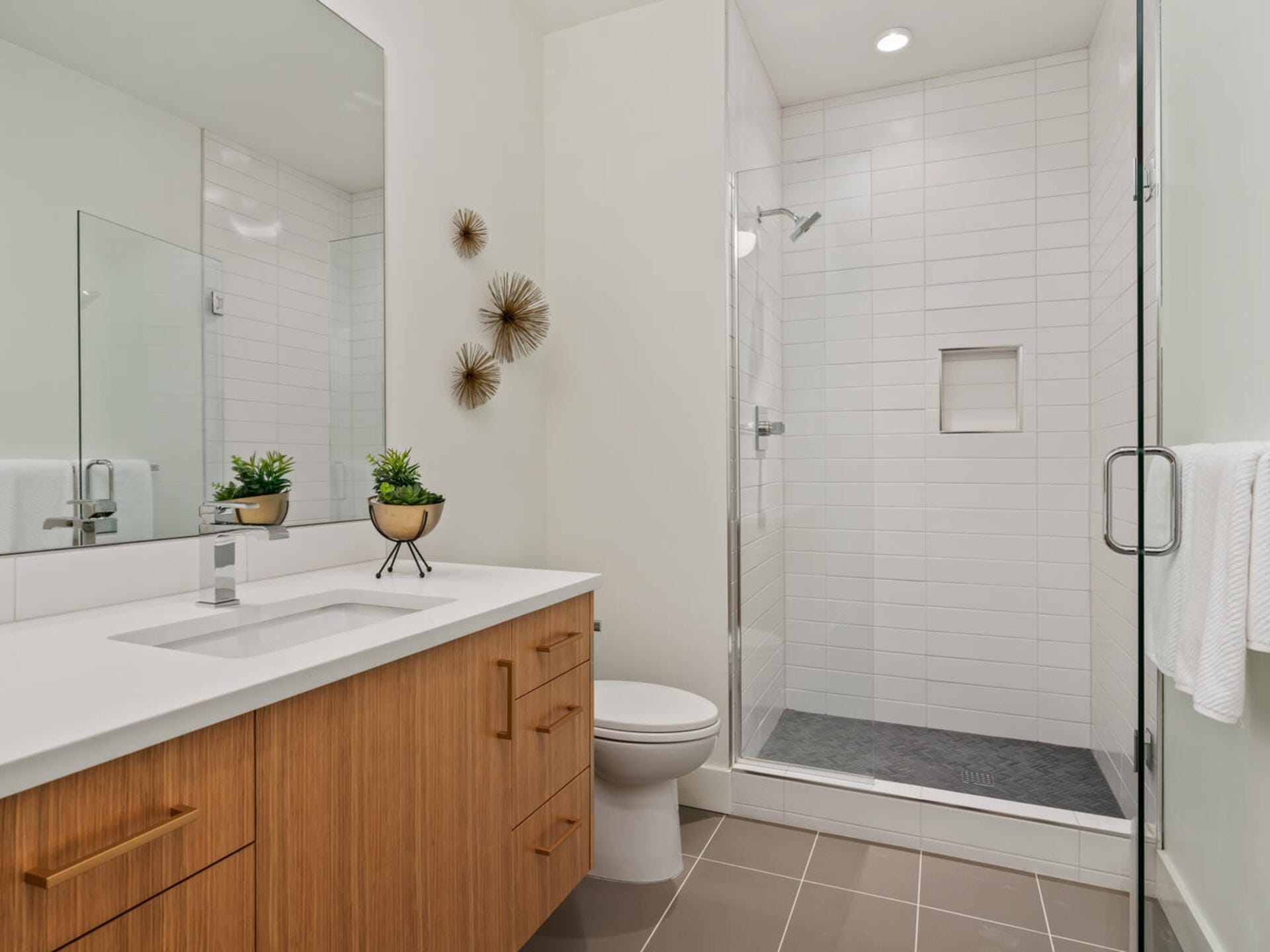 Modern bathroom featuring a wooden vanity with a white countertop, sink, and two potted plants. A glass-enclosed shower with white tiles stands in the background. Two decorative wall pieces are mounted on the wall above the toilet.