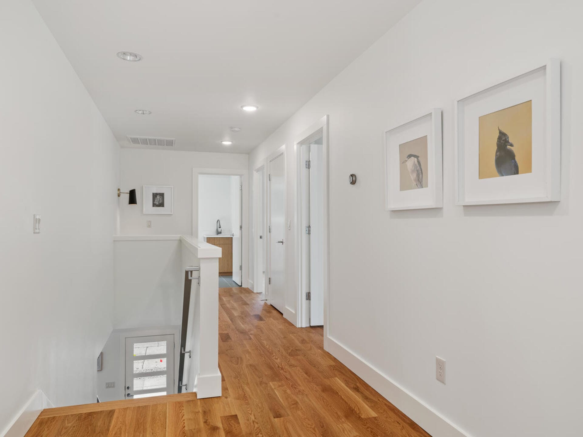 A hallway with white walls and wooden floors. Three closed doors are along the right wall, which has two framed bird illustrations. At the end of the hallway, theres a glimpse of a room with a small cabinet and artwork. A staircase is on the left.