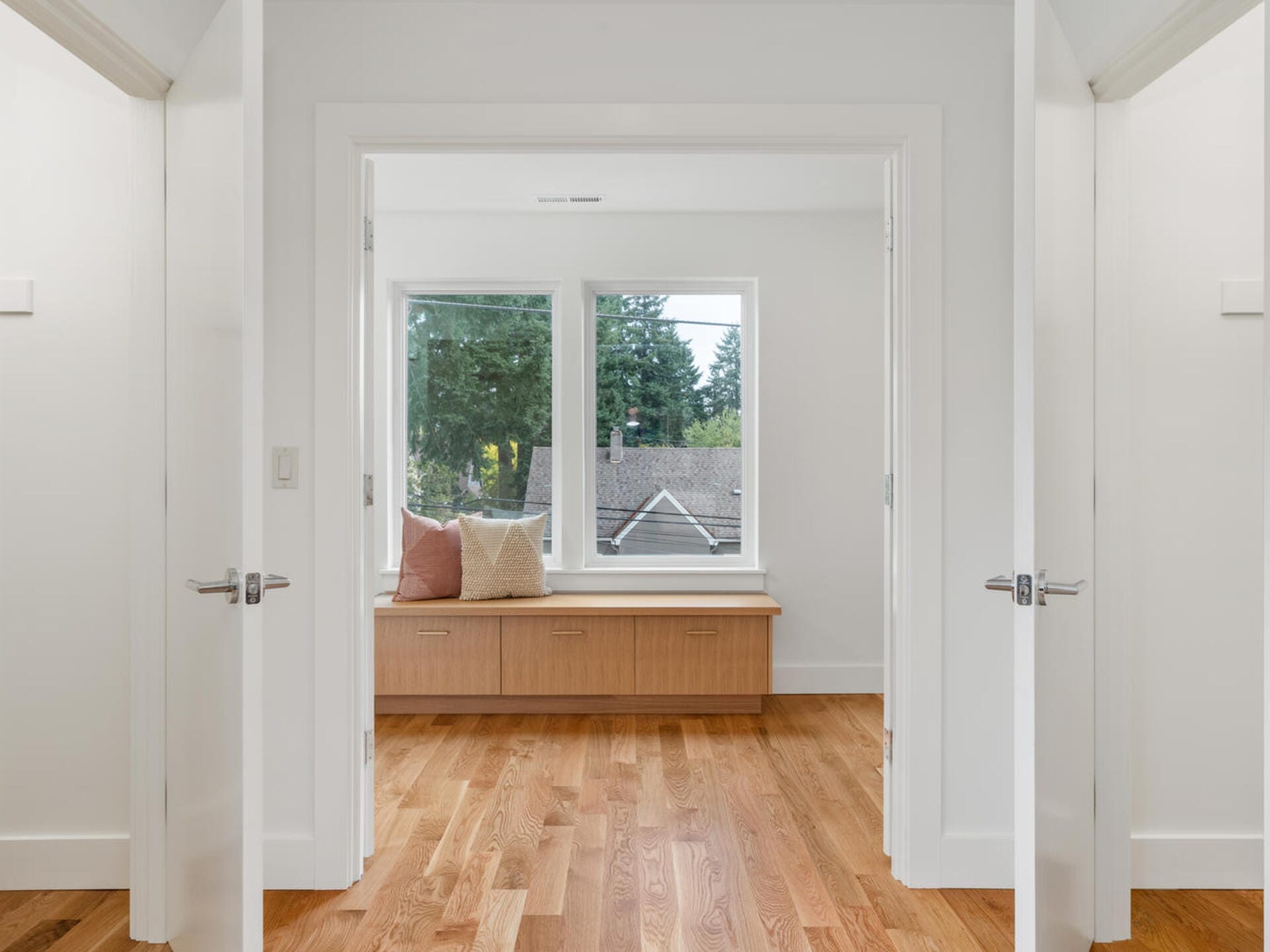 Bright room with wooden floors and white walls, featuring a window seat with cushions. Large windows offer a view of trees and nearby rooftops. Double doors frame the entrance, enhancing the open and airy feel.