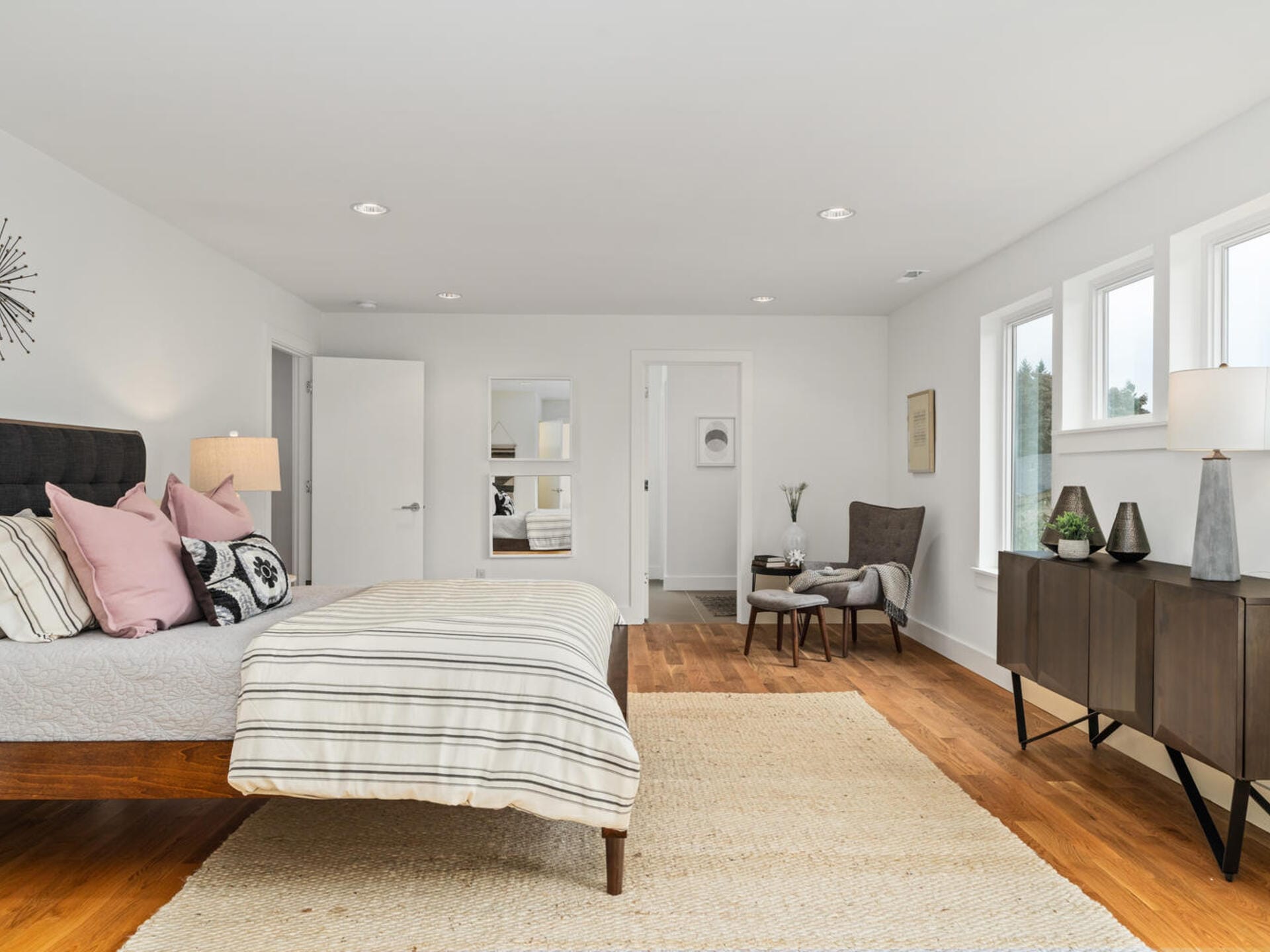 A spacious, modern bedroom with a large bed featuring striped bedding and pink pillows. A wooden floor and light-colored walls enhance the minimalist decor. A brown chair and a small table sit by the window, and a woven rug covers part of the floor.