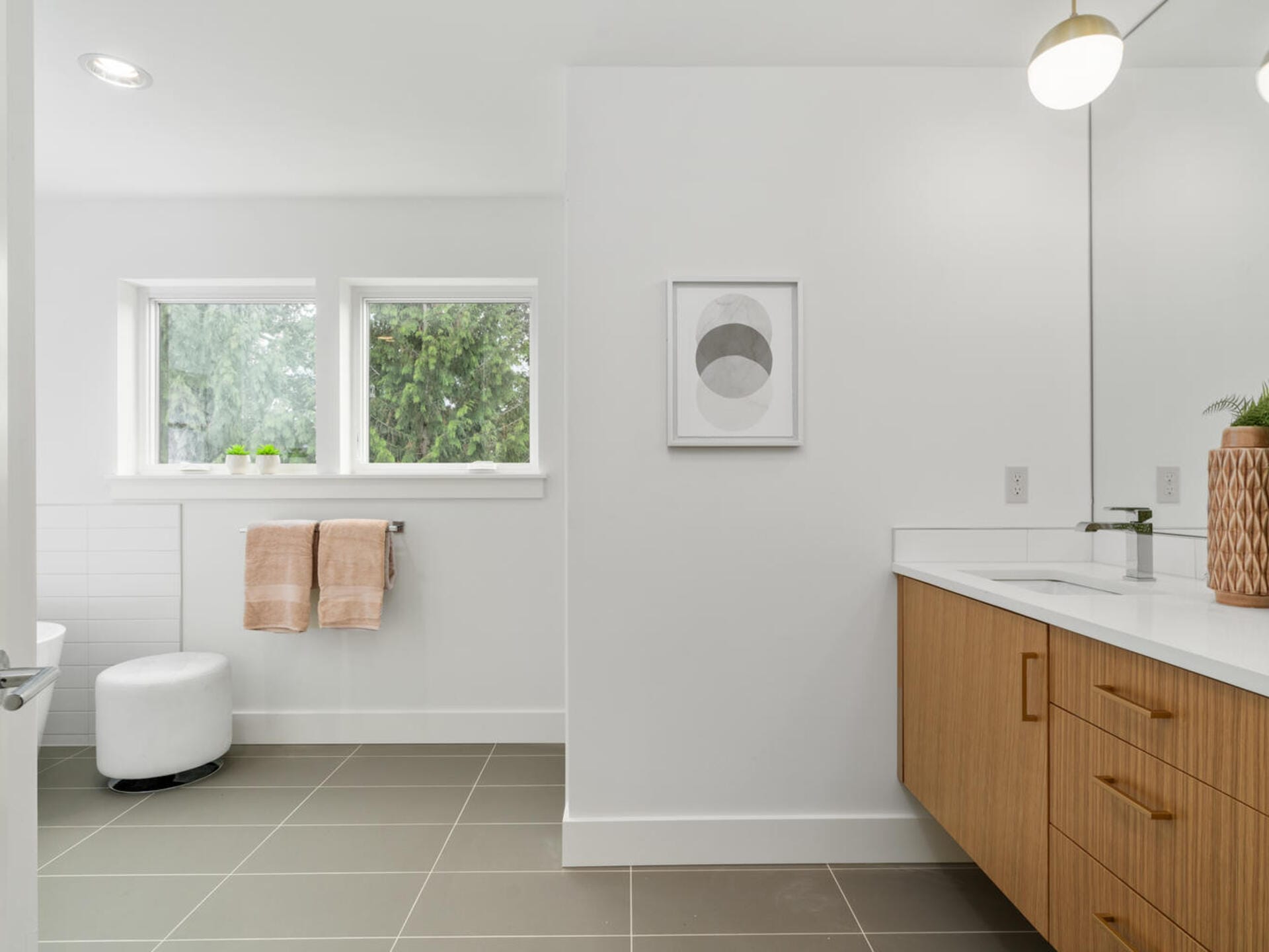 Modern bathroom with a wooden vanity and white countertop. A mirror with two round lights is above the vanity. Two large windows let in natural light. A framed abstract art piece adorns the wall. Towels hang by the window, and a small stool is present.