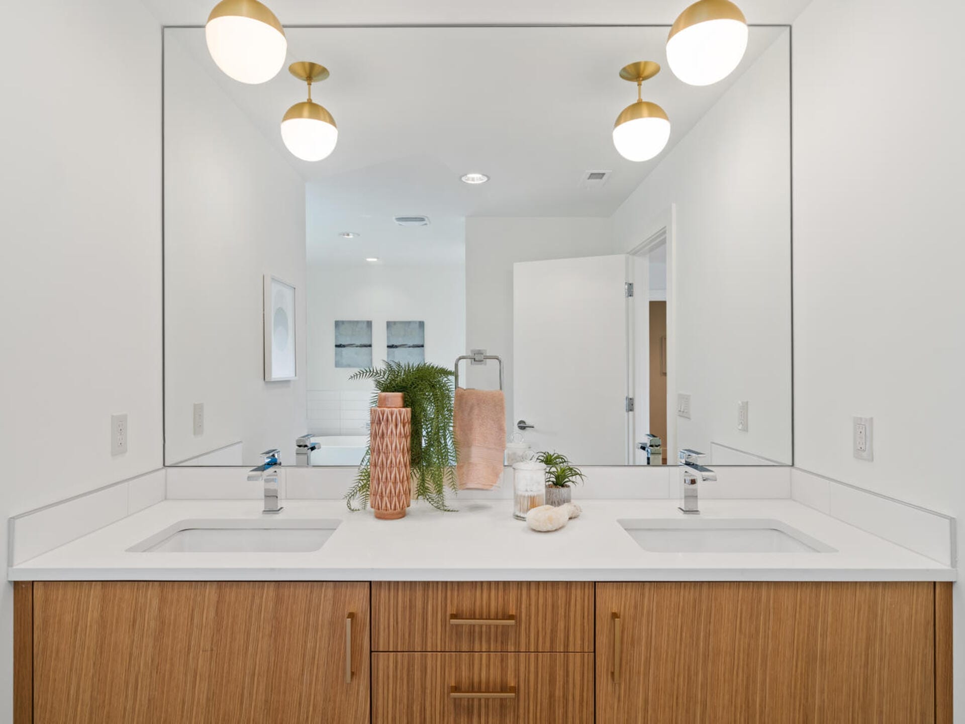 A modern bathroom with a double sink vanity featuring a large mirror. The countertop is white with a vase containing green plants and a folded towel. The room is well-lit by three ceiling lights, and an open door is visible in the reflection.