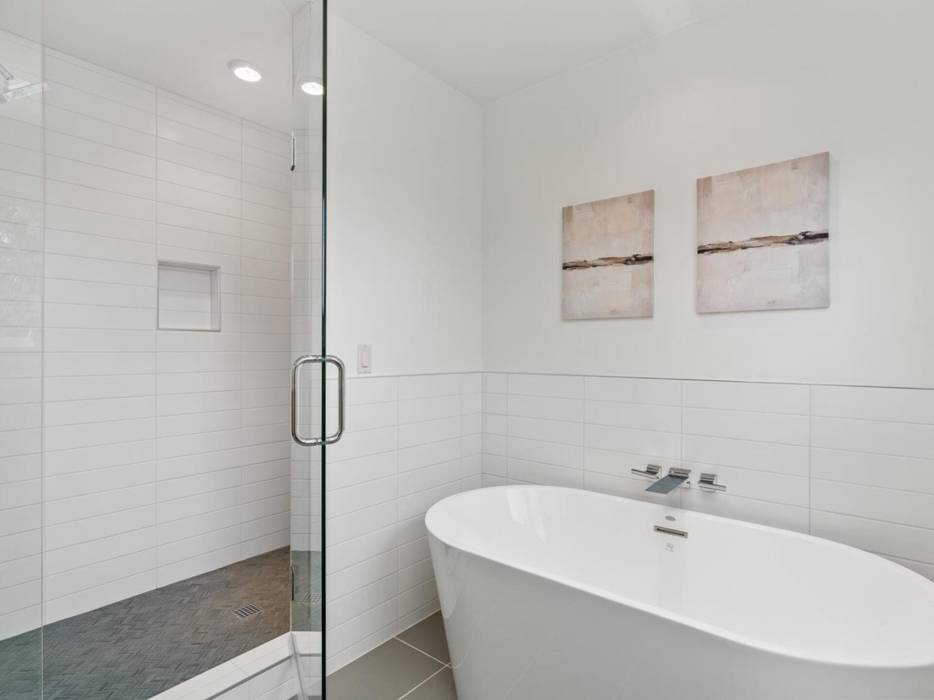 Modern bathroom with a white freestanding bathtub next to a glass-enclosed shower. White tiles line the walls, and two abstract paintings hang above the tub. The floor is covered with dark tiles.