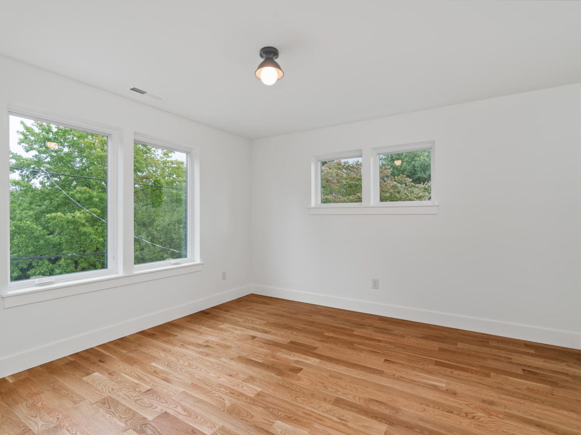 An empty room with white walls and a wooden floor. There is a ceiling light fixture and two large windows on the left wall, as well as two smaller windows on the adjacent wall, offering views of green trees outside.