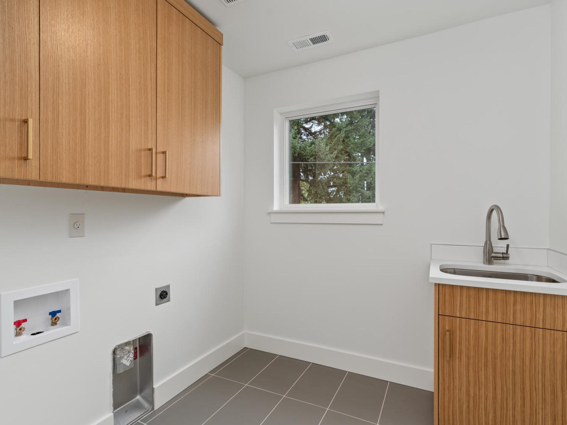 A modern laundry room with a wooden cabinet, a small sink with a faucet, and a window. The walls are white, and the floor is tiled in gray. There are connections for a washer and dryer on the left wall.