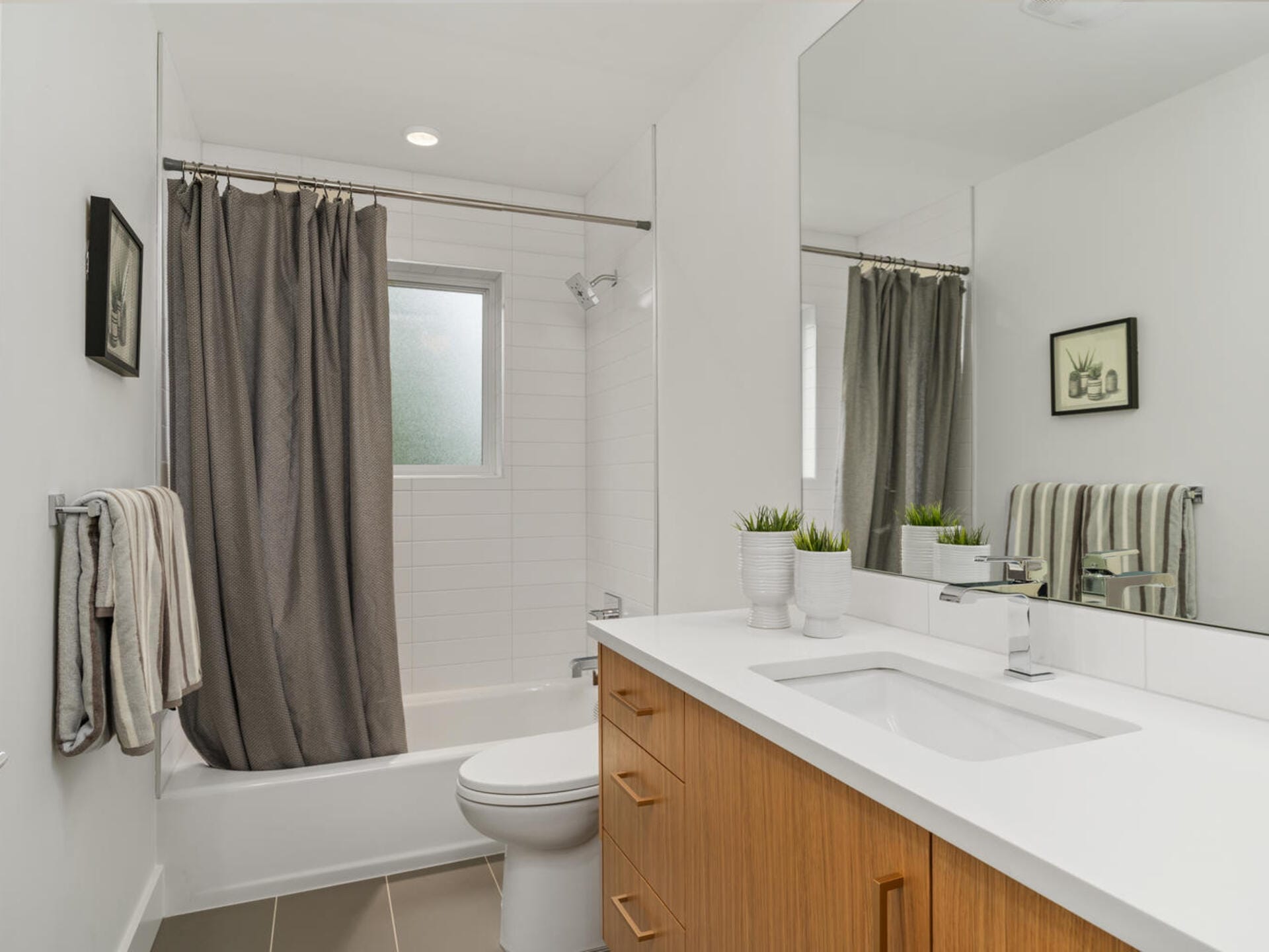 A modern bathroom with a large mirror, white countertop, wooden cabinet, and a sink. A toilet and towel rack with a hanging towel are beside a bathtub with a gray shower curtain. A small window and wall art complete the decor.