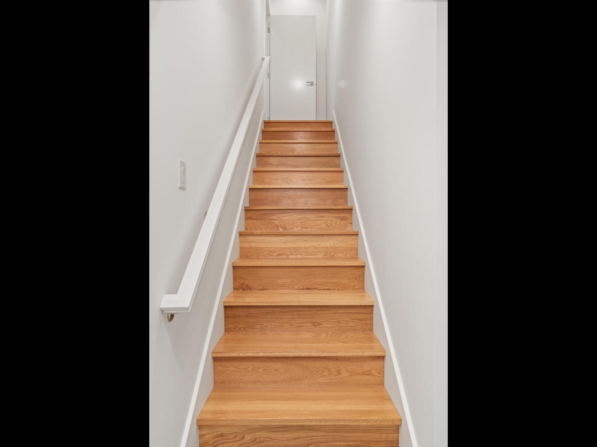 A wooden staircase leading upwards in a narrow hallway with white walls. A handrail is fixed on the left wall. At the top, there is a closed white door. The space is well-lit from above.