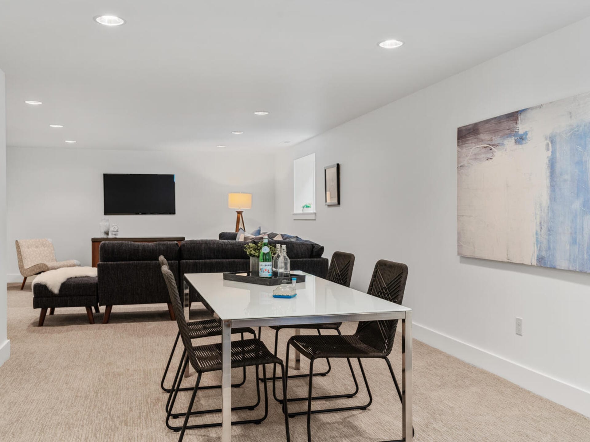 A modern living room with a sleek white dining table, six black chairs, and a dark grey sofa. A large flat-screen TV is mounted on the wall. Art decorates the walls, and a cozy chair with a throw blanket completes the space.
