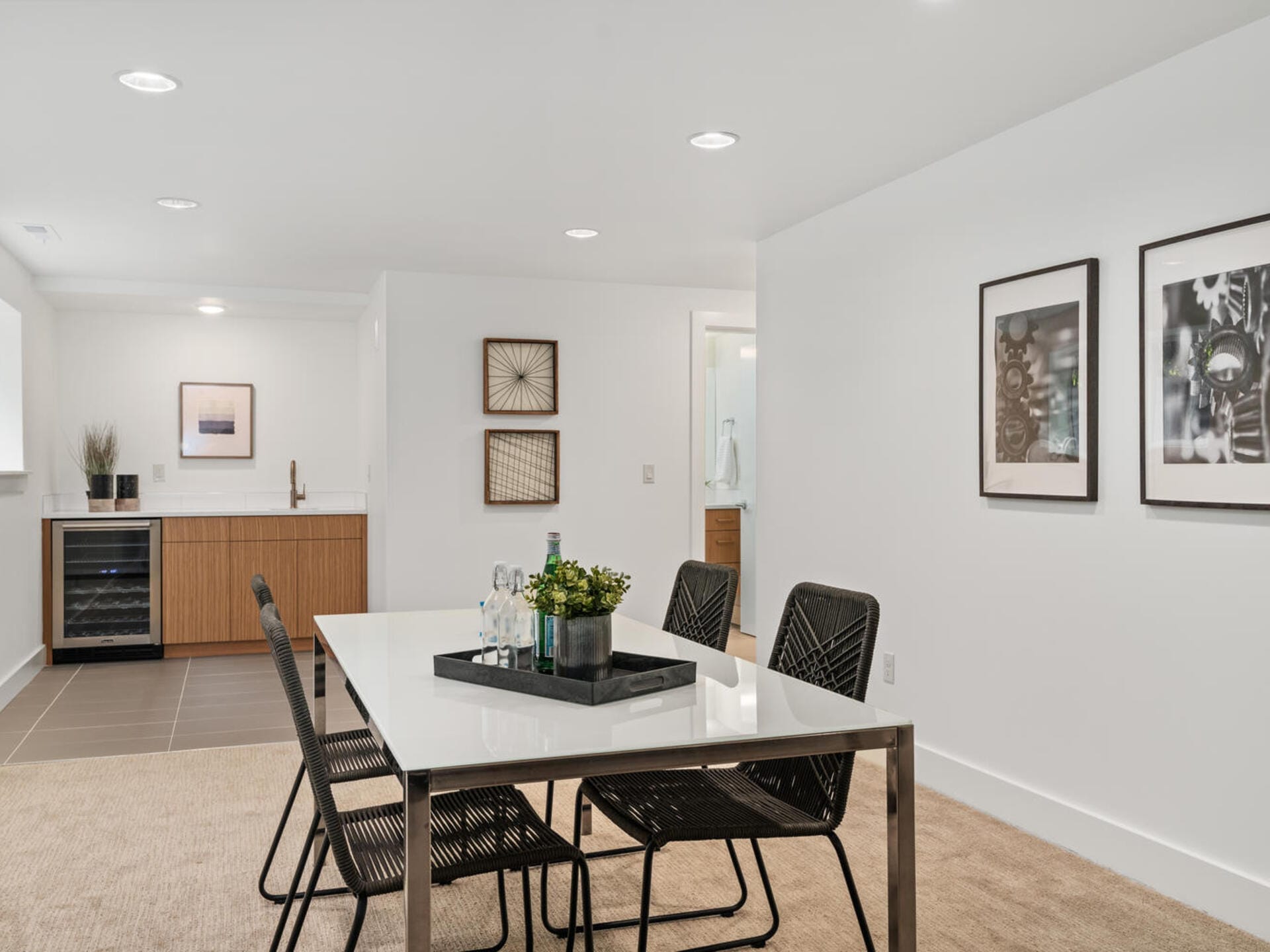 A modern dining area with a sleek white table and four black mesh chairs. The room features minimalist decor with framed art, a small kitchenette with a wooden cabinet, wine cooler, and recessed lighting. Neutral tones create a clean and cozy atmosphere.