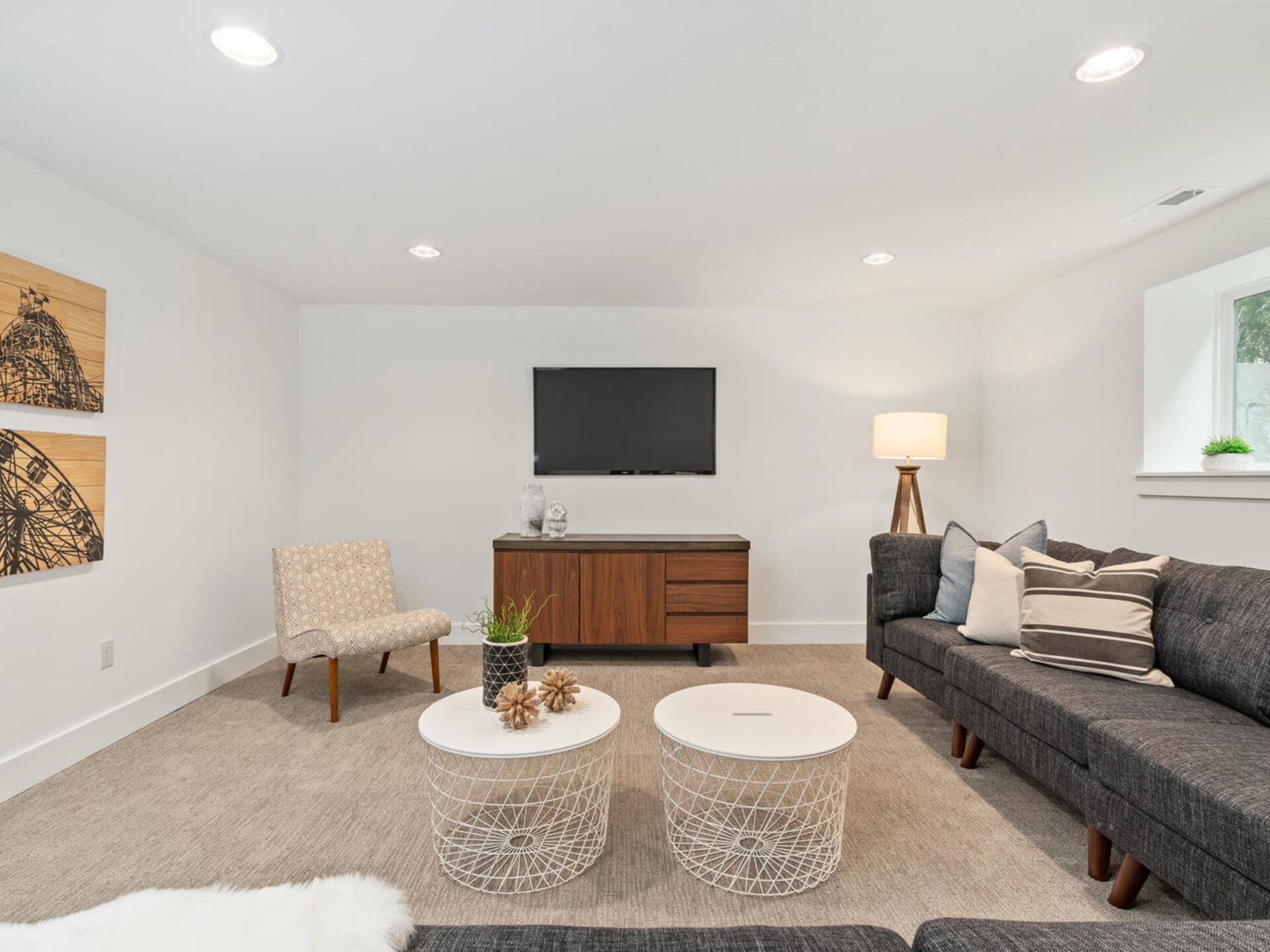 A modern living room with beige carpet, dark gray sectional sofa, patterned armchair, and two white round coffee tables. A wooden media console with a TV is against a white wall, under three framed art pieces. A floor lamp stands beside the sofa.
