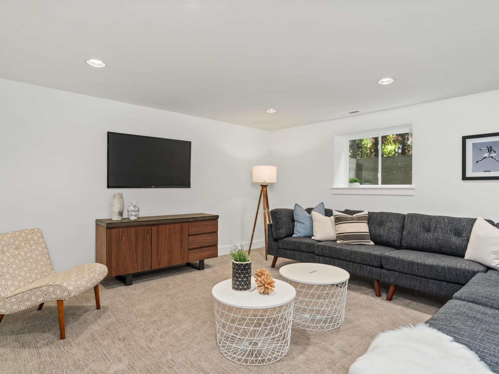 A modern living room with a gray sectional sofa, patterned chair, two white round coffee tables, and a wooden media console with a mounted TV. A tall floor lamp and a window provide light, and a framed picture is on the wall.