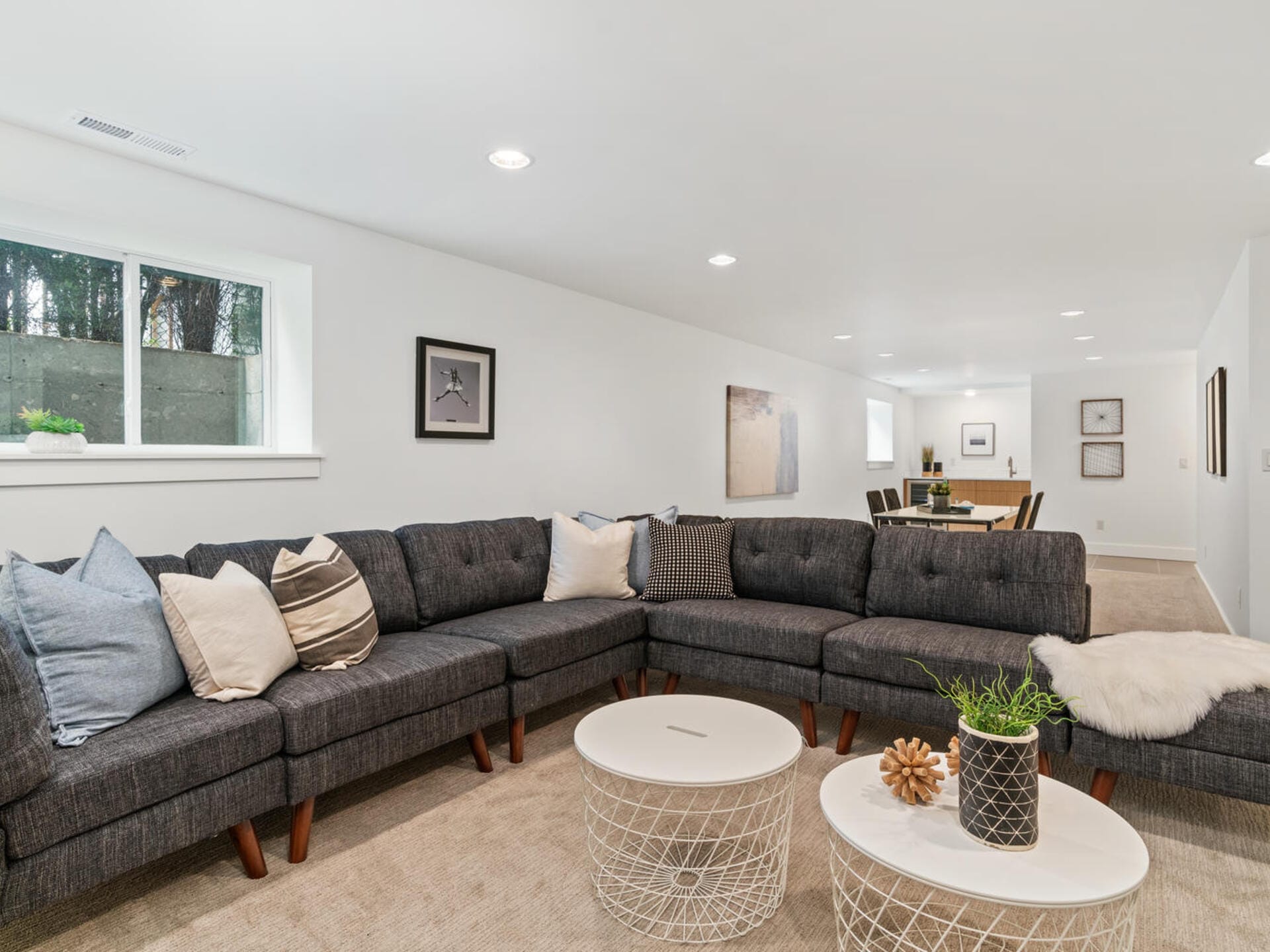 A modern living room with a large dark gray sectional sofa adorned with various pillows. Two round white coffee tables sit in front, one with a plant. The room features minimalist decor, framed artwork, and a dining area in the background.