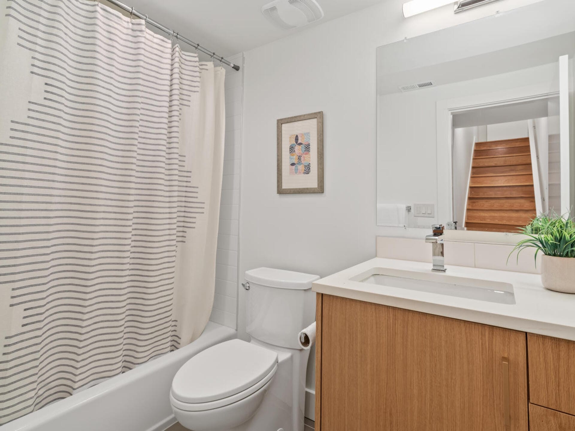Bathroom with a white toilet and a wooden vanity featuring a white sink. A shower with a beige and gray-striped curtain is on the left. A plant is on the counter, and framed art hangs above the toilet. Wooden stairs are visible in a mirror.
