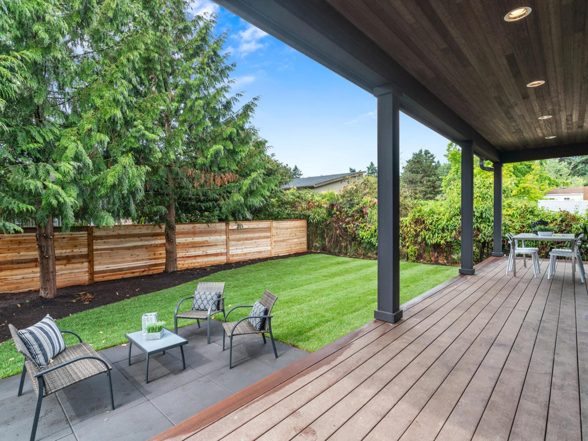 A backyard with a wooden deck and seating area featuring four chairs and a small table on a paved surface. The yard has a well-maintained lawn, a wooden fence, and surrounding greenery, including trees and shrubs, under a partly cloudy sky.