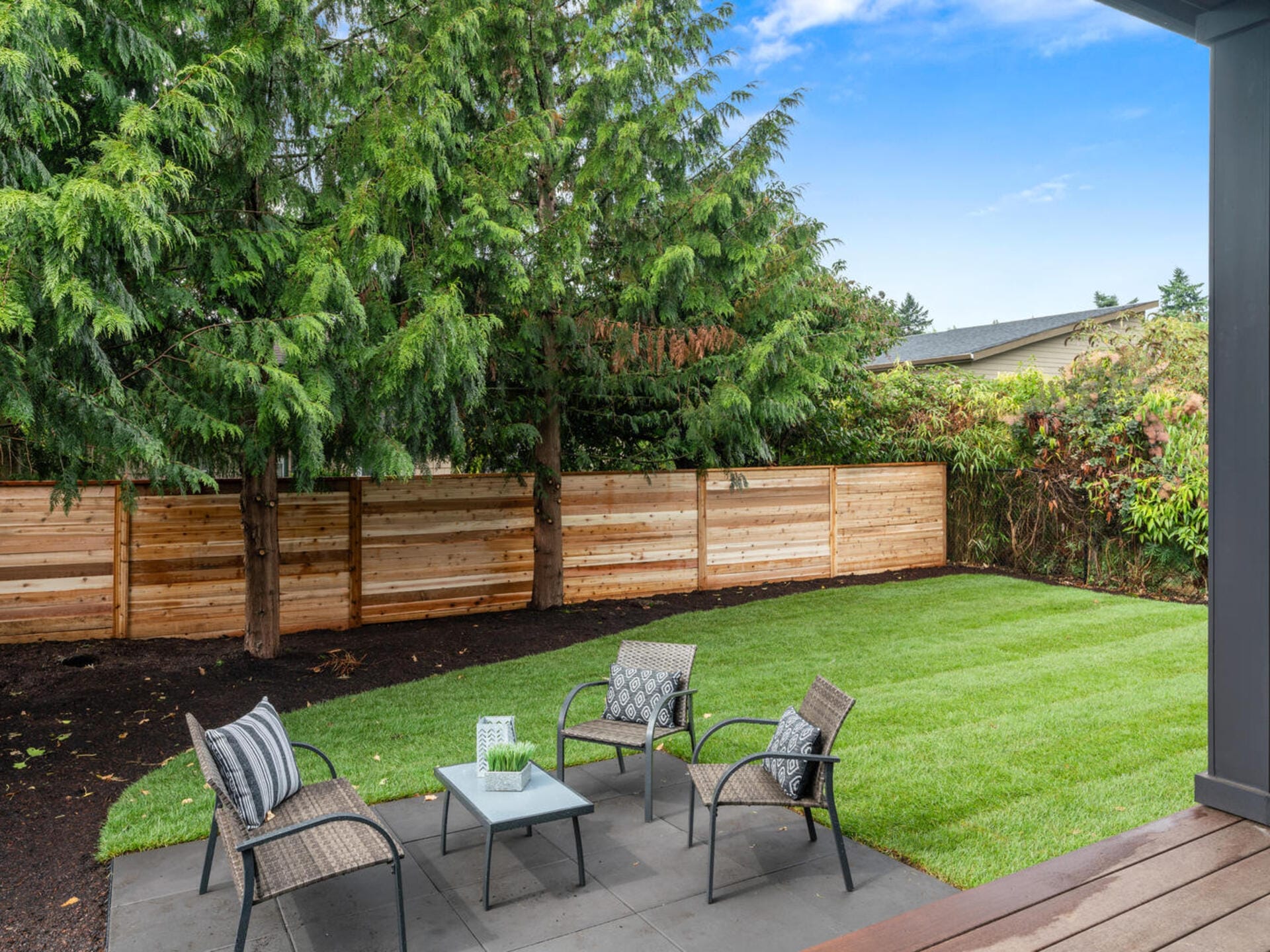 A backyard with a grassy lawn, a wooden fence, and several tall trees. There is a small patio area with four chairs around a glass-topped coffee table, all embellished with patterned cushions. The sky is clear and blue.