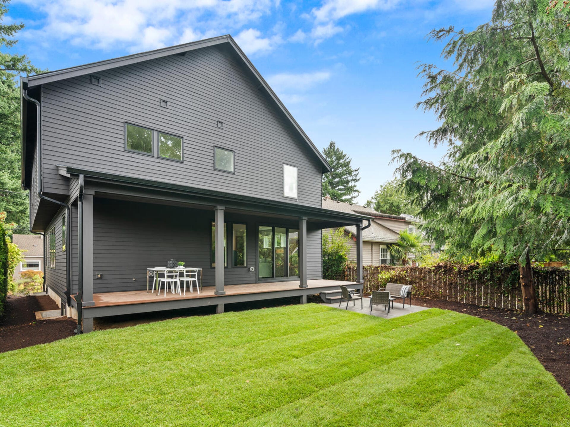 A modern house with dark gray siding, large windows, and a covered patio. The backyard features a green lawn, outdoor seating on the patio, and surrounding trees. The sky is clear with a few clouds.