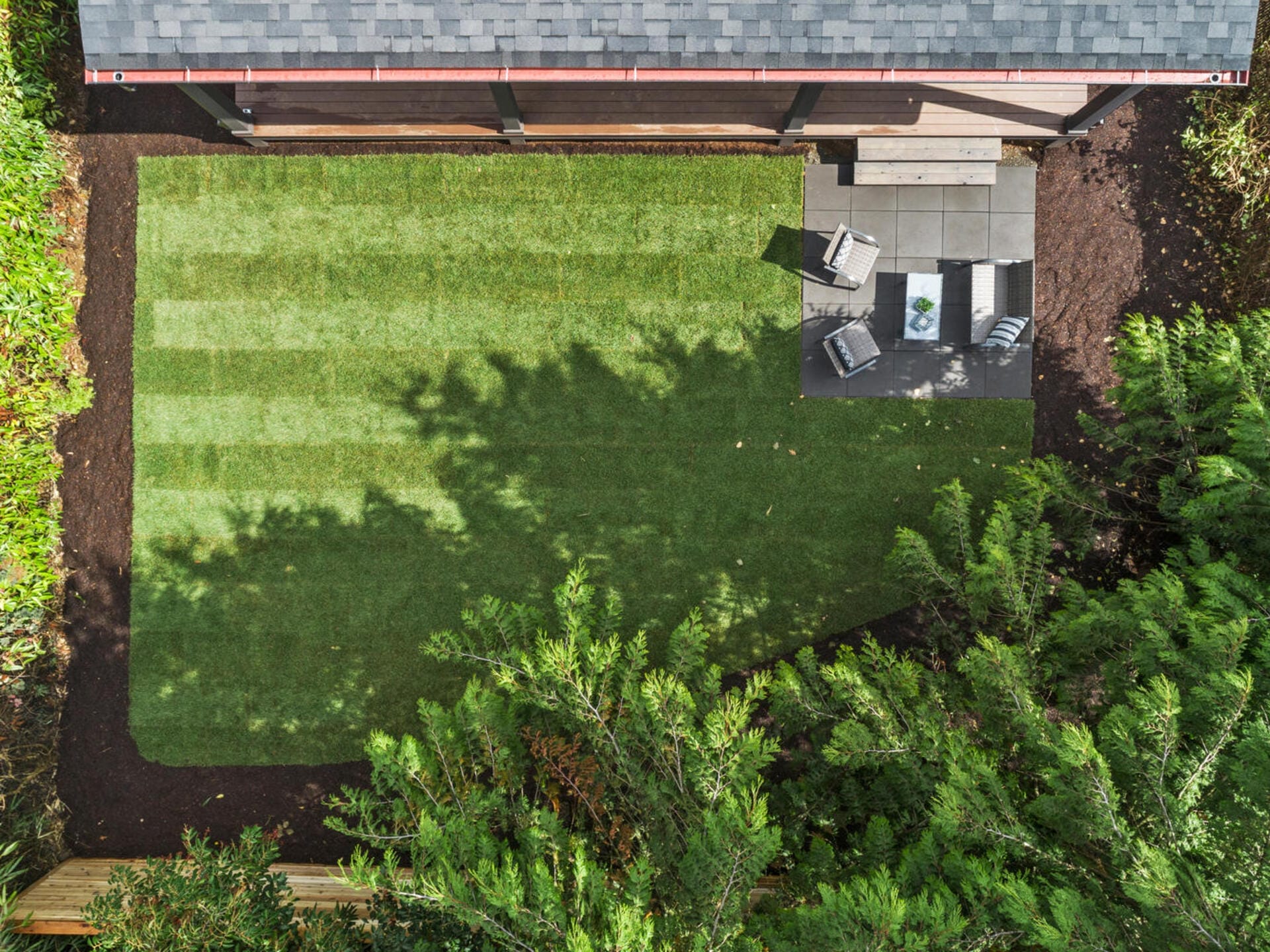 Aerial view of a small backyard with a neatly mowed lawn and a patio area. The patio has outdoor furniture set up for seating, and its adjacent to a garden shed. Surrounding the yard are trees and shrubs, providing a lush, green border.