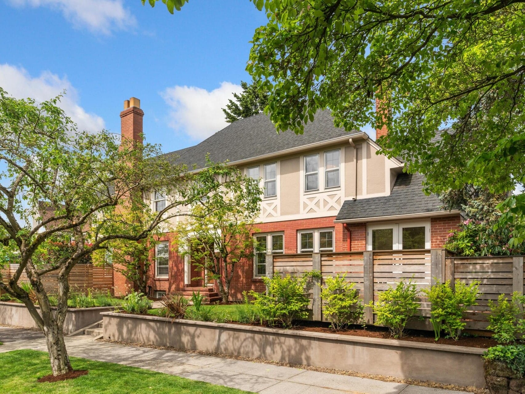 A two-story brick and stucco house with a chimney, surrounded by a wooden fence. The front yard features a well-maintained garden with trees and shrubs. The sky is clear and blue, enhancing the homes inviting appearance.