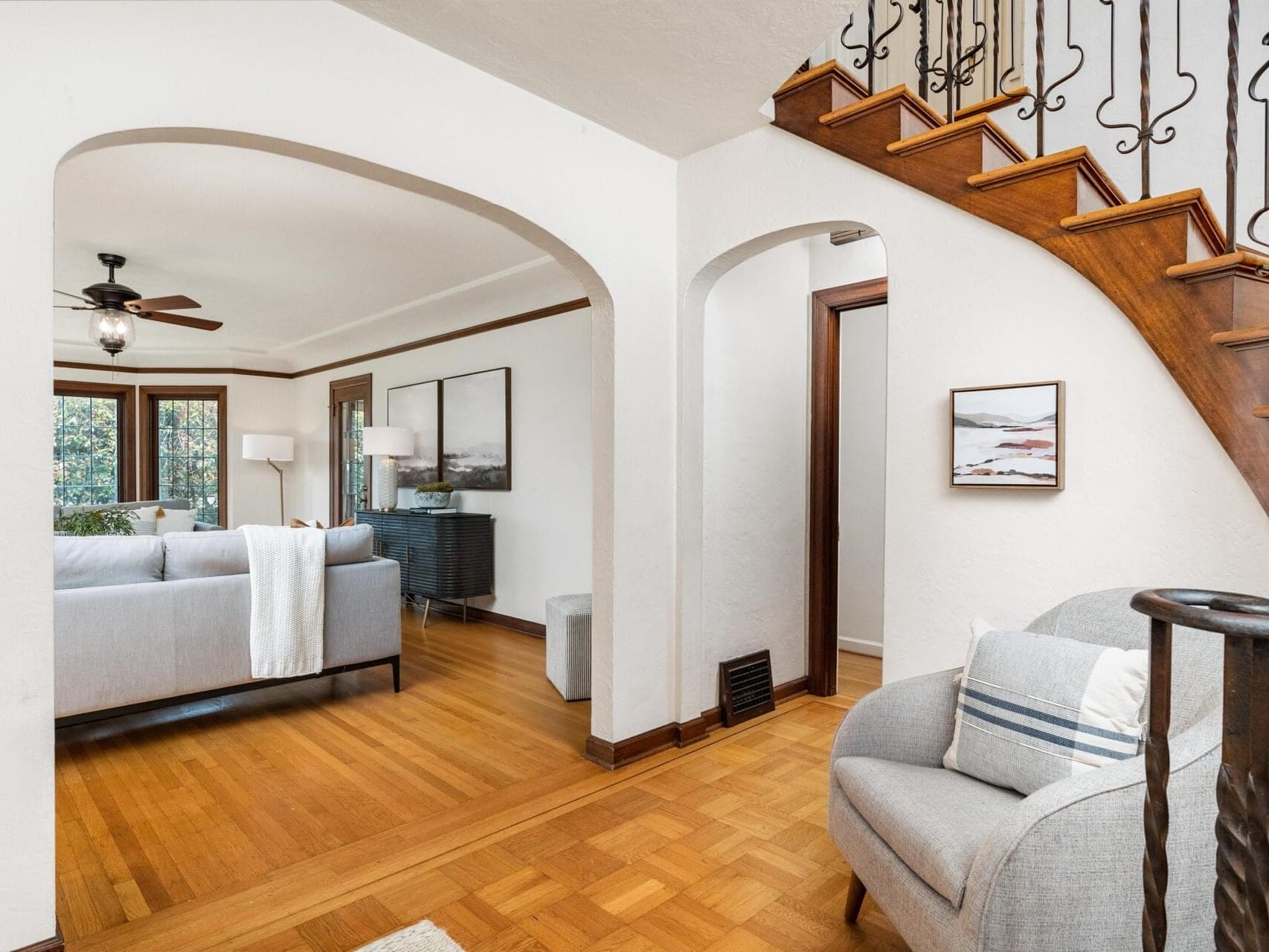 Interior of a modern living room with wooden floors, a gray sofa, and matching armchair. An archway leads to a sitting area with two chairs and artwork. A wooden staircase with iron railing is visible on the right. Ceiling fan and natural light included.