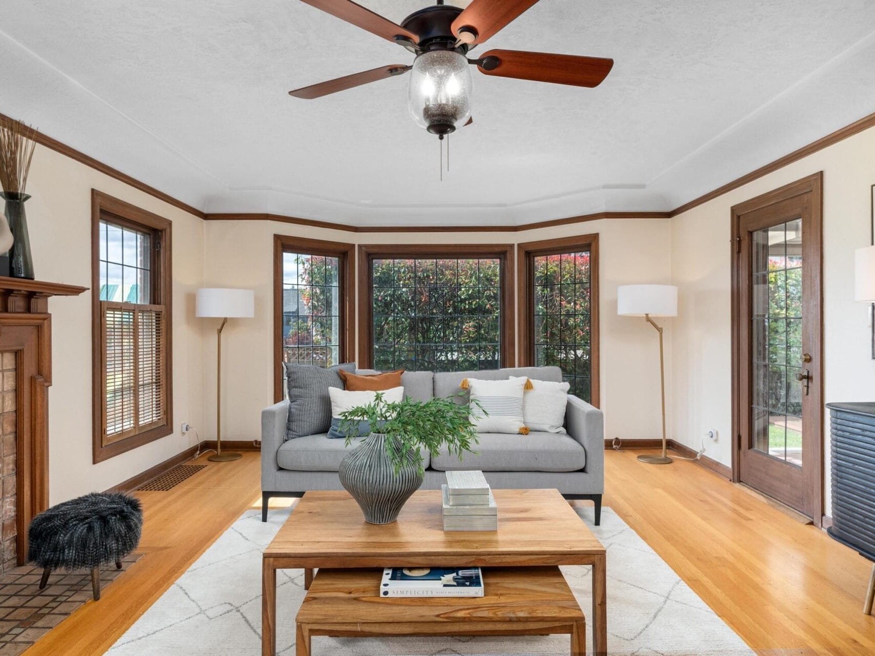 A cozy living room with a gray sofa, a wooden coffee table, and a decorative plant. The room features wooden floors, a ceiling fan, and a fireplace. Large windows and a glass door let in natural light, enhancing the rooms warm ambiance.