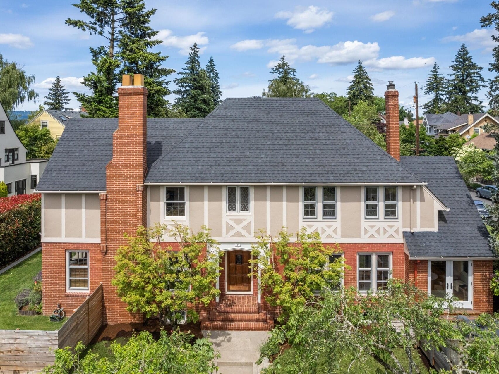 An elegant two-story house with a dark gray roof and red brick facade, featuring large windows and lush greenery in the front yard. Tall trees and neighboring homes are visible under a clear blue sky.