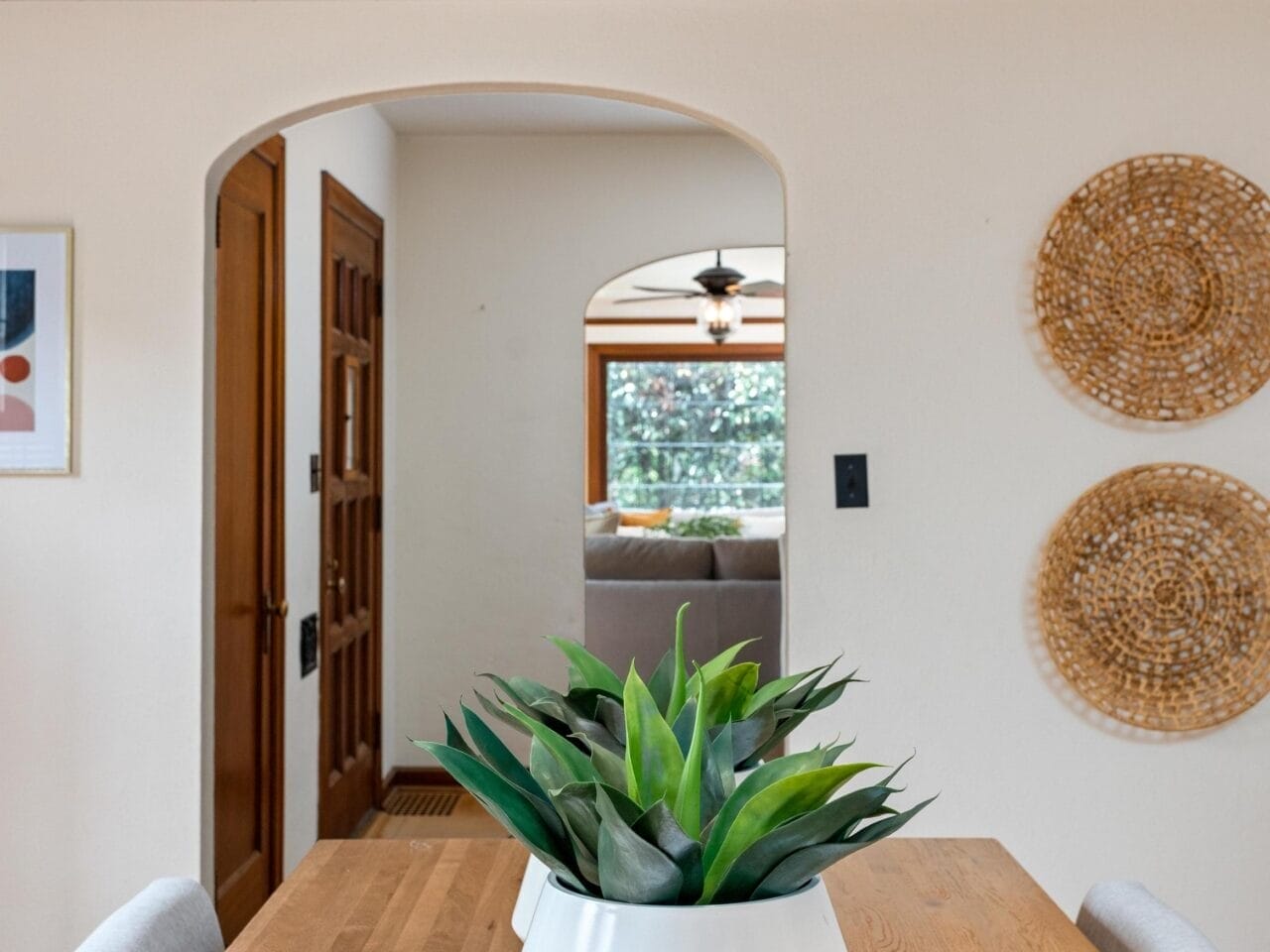 A dining area with a wooden table and a white planter holding green plants. A large white lamp hangs above. The room leads to a living area with a couch, visible through an arched doorway, and woven wall decor on the side walls.