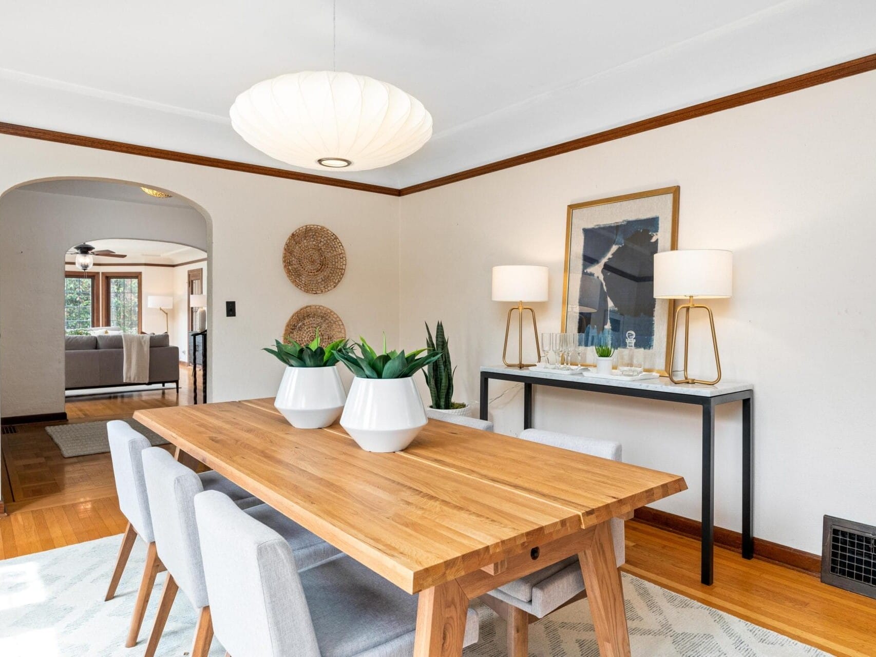 A bright dining room with a wooden table and six gray chairs. Two white planters sit on the table. A console with two lamps and a framed art piece is in the background. The room features arched doorways and wooden accents.