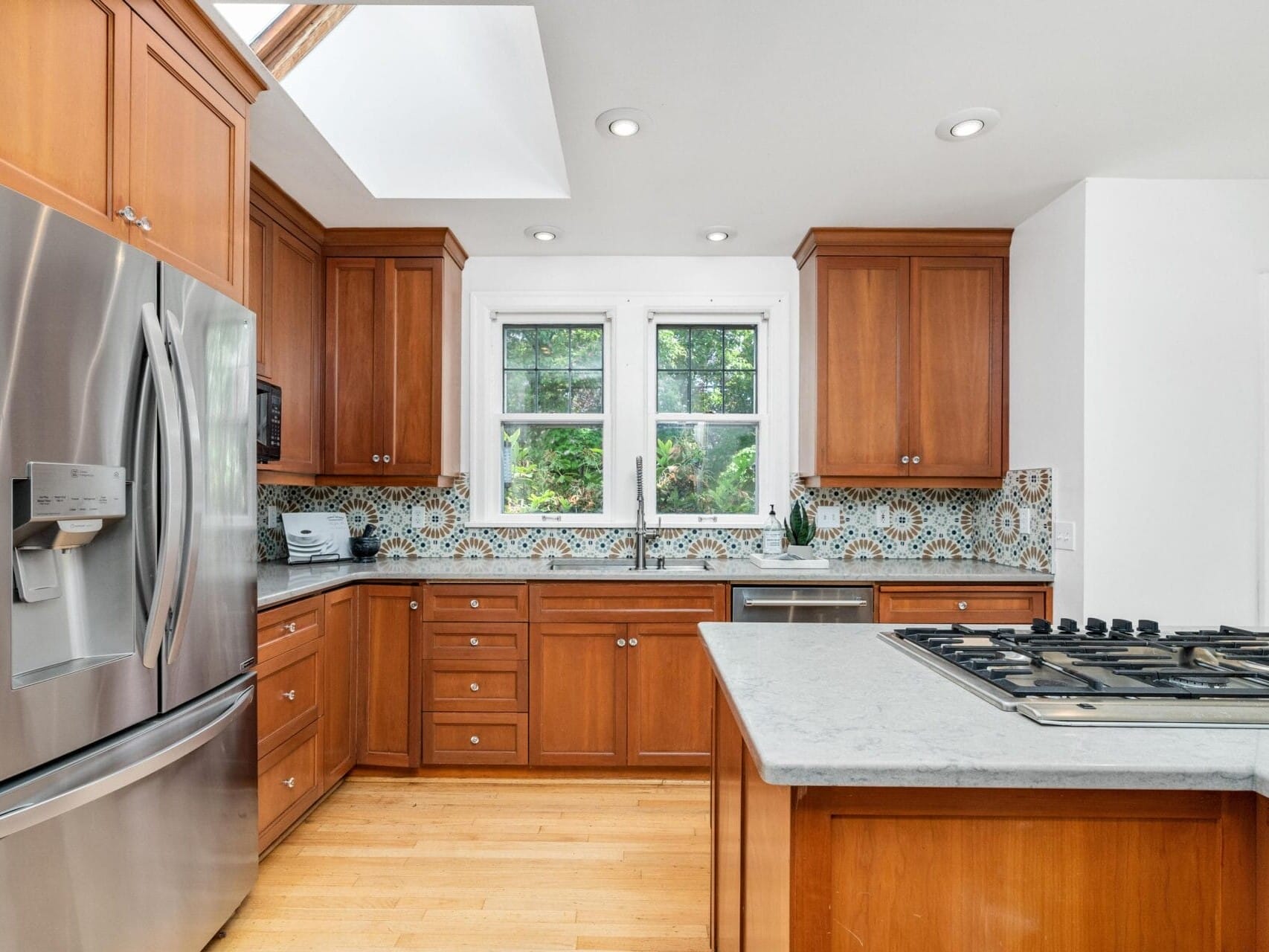 A modern kitchen with wooden cabinets, a stainless steel refrigerator, and a gas stovetop on a central island. There are patterned tiles as a backsplash and two large windows providing natural light.