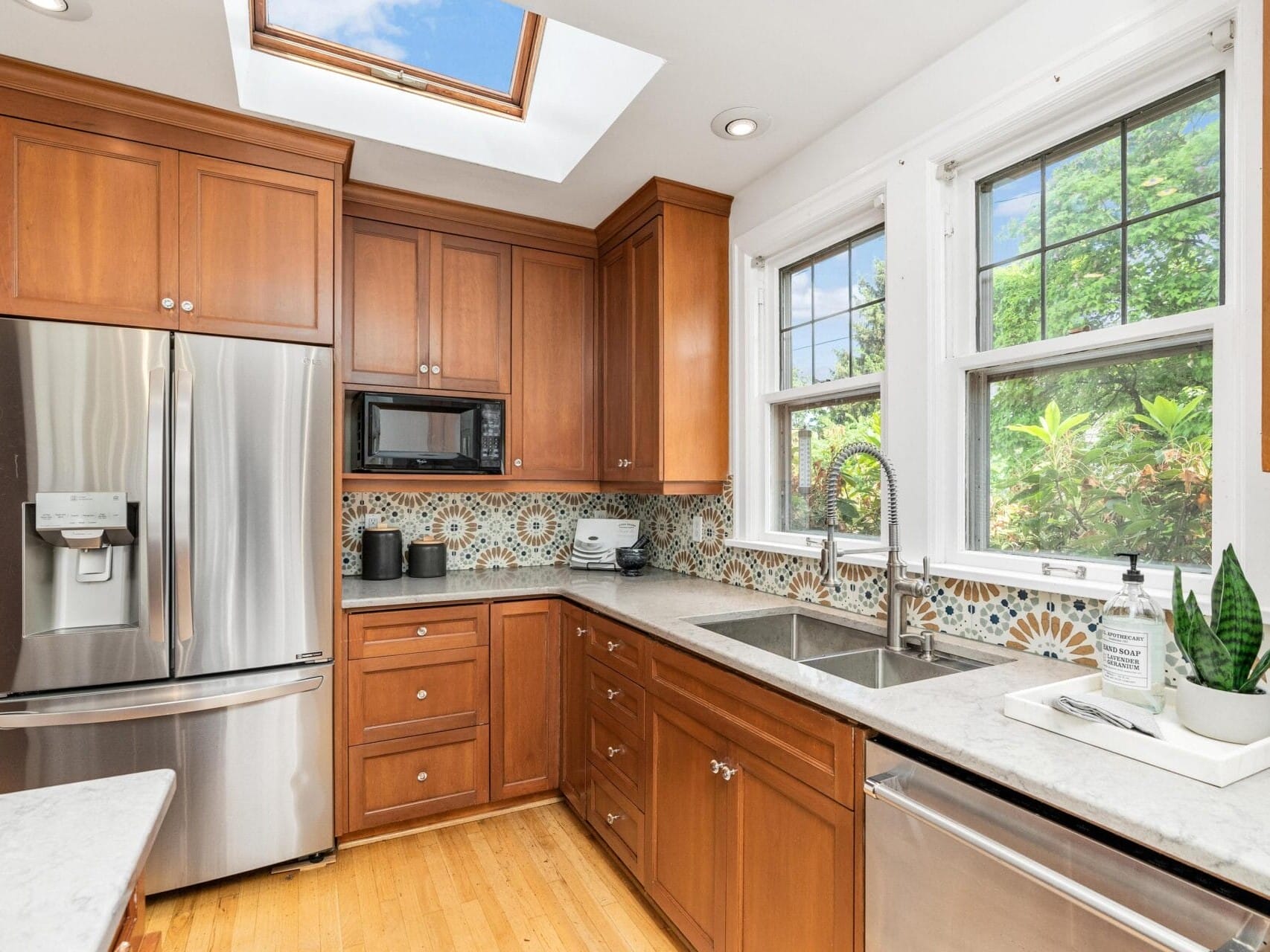 A kitchen with wooden cabinets, a stainless steel fridge, and a microwave. There are two sinks with a floral backsplash design. Large windows provide natural light, and plants are visible outside. A skylight is above, illuminating the space.