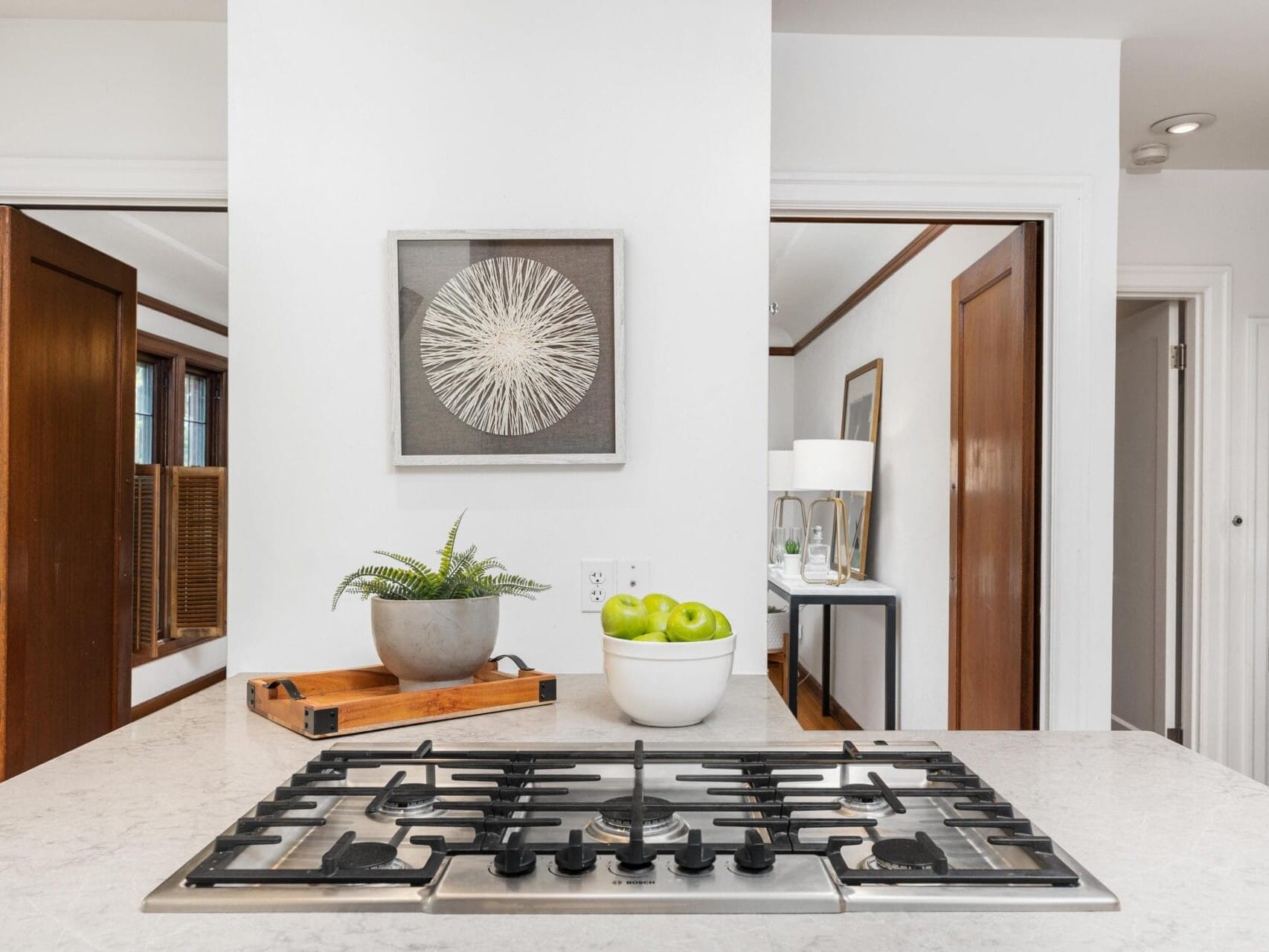 A modern kitchen with a gas stovetop on a white countertop. A decorative plant and a bowl of green apples sit to the left. A framed circular artwork hangs on the white wall. Wood doors and a glimpse of a hallway are visible in the background.