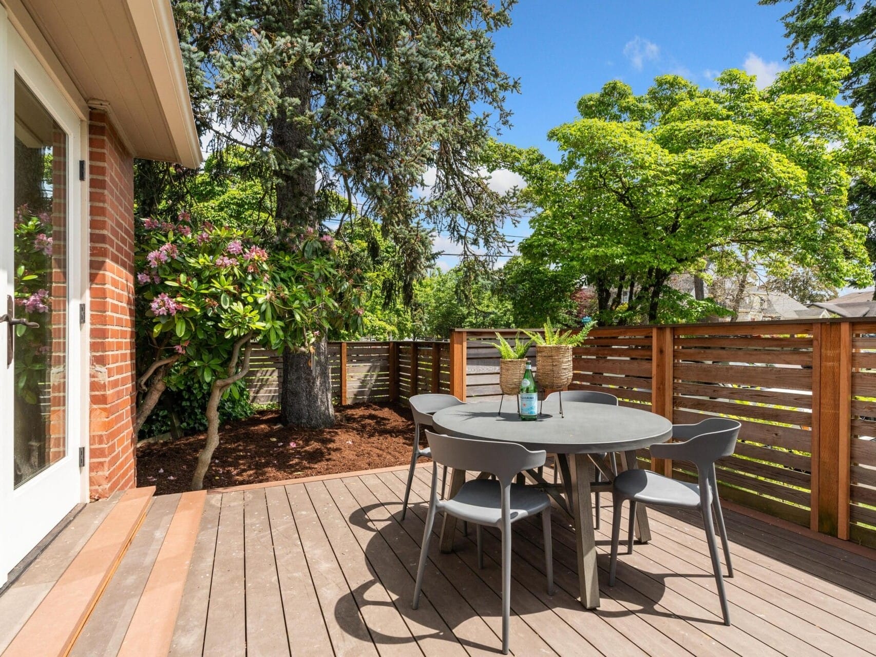 A wooden deck surrounded by a garden features a round gray table with four matching chairs. The table has a bottle of wine and glasses. A wooden fence and lush green trees provide a scenic backdrop under a clear blue sky.