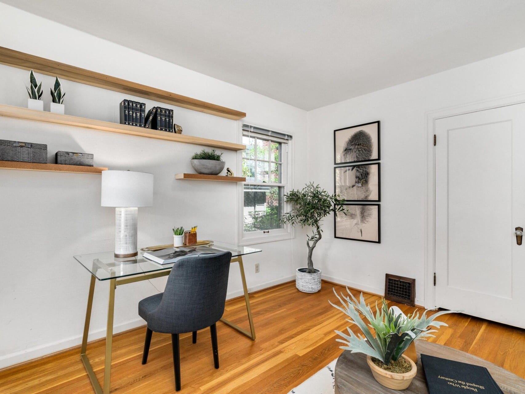 A bright home office with a glass desk, black chair, and potted plants. Wooden shelves hold decor items and three framed pictures are on the wall. A lamp and a book are on the desk, and a window lets in natural light. Hardwood floor.