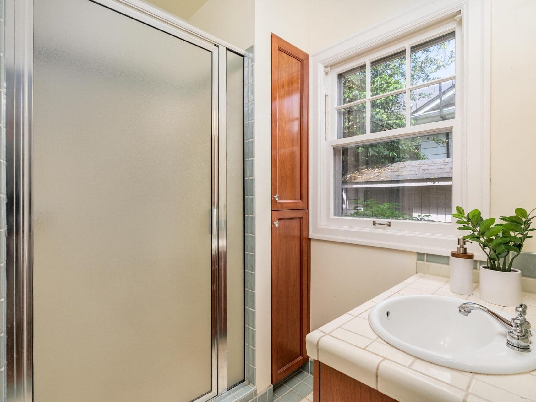 A bathroom with a frosted glass shower door, a wooden cabinet, and a window. A white sink is set in a tiled countertop with two small potted plants. Natural light streams in through the window.