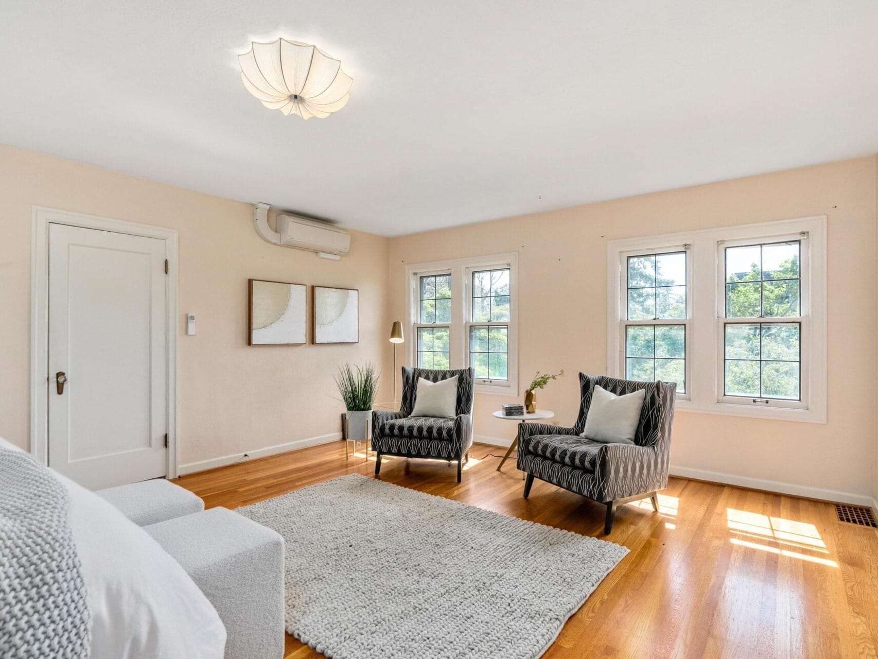 A bright living room with wood flooring, white walls, and a large gray area rug. It features two gray armchairs, a small side table, a white sofa, and a ceiling light. Three windows let in natural light, and two framed artworks hang on the wall.