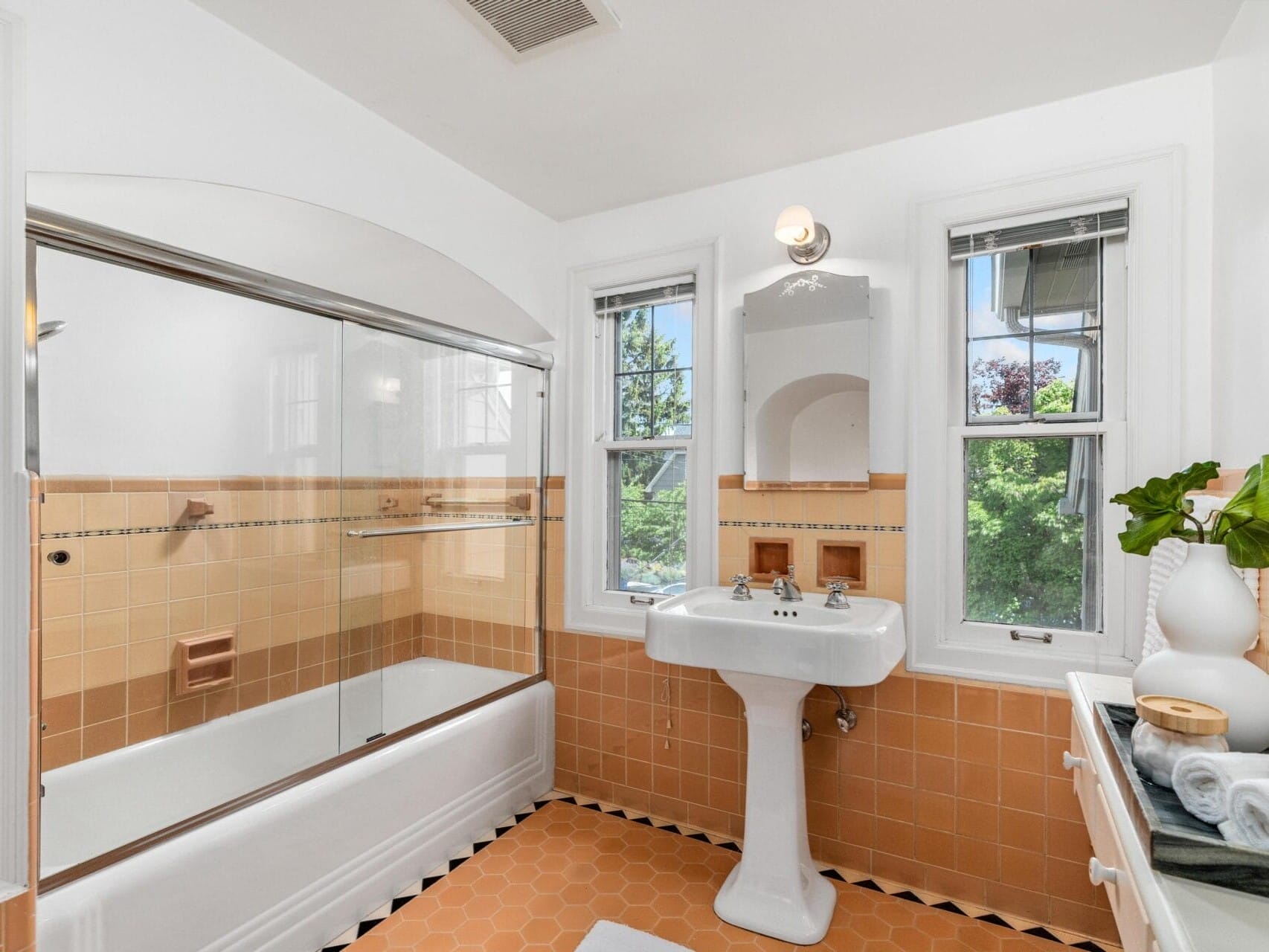 A vintage-style bathroom with peach-colored tiles features a pedestal sink and a bathtub with a sliding glass door. Two windows provide natural light, and a small cabinet holds a plant and toiletries. A single light fixture is mounted above the mirror.